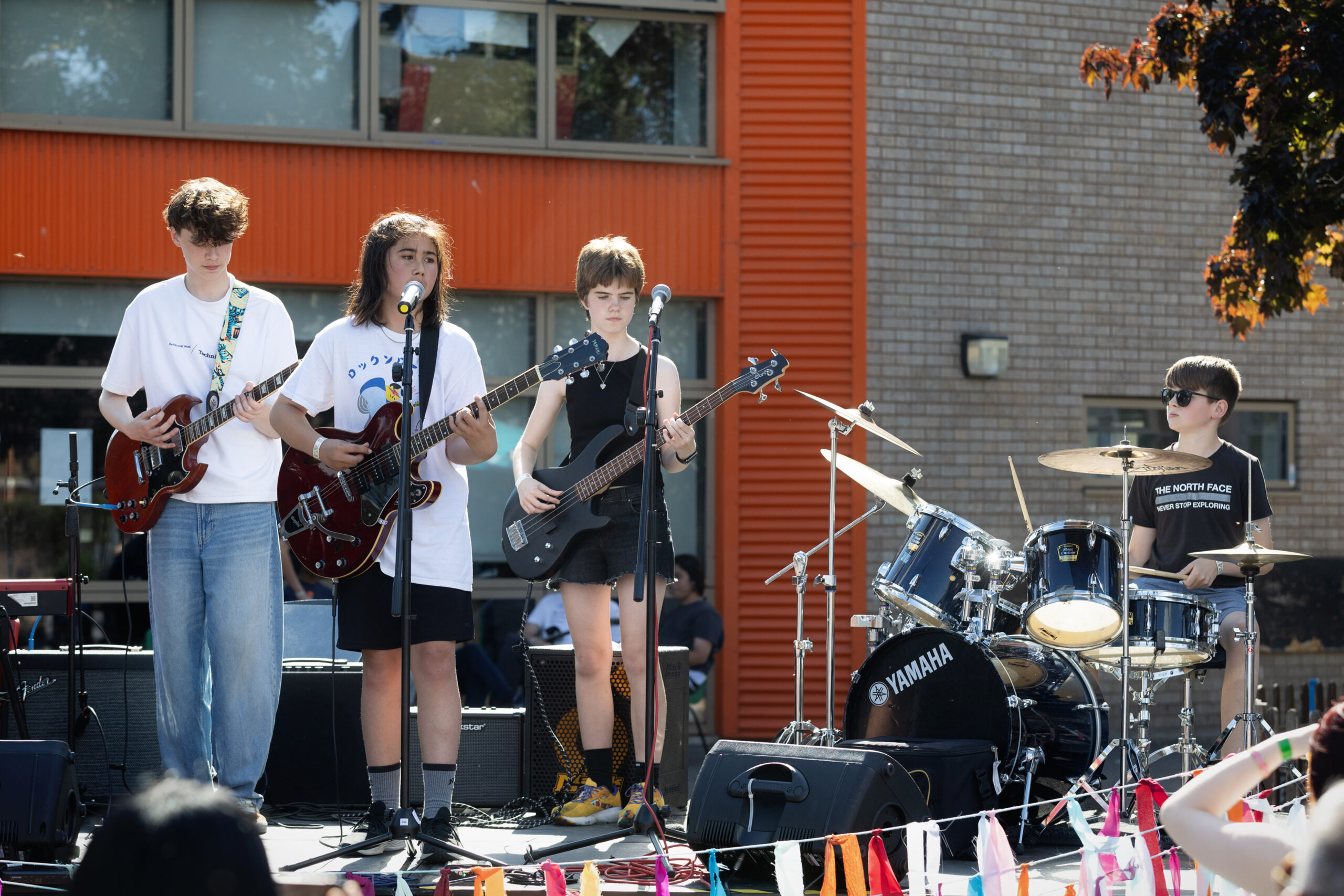 A youth band amplifies their sound outdoors on a stage. Four kids play electric guitar, bass, and drums, while one sings into a microphone. Colorful flags decorate the stage, with a brick building and tree in the background.