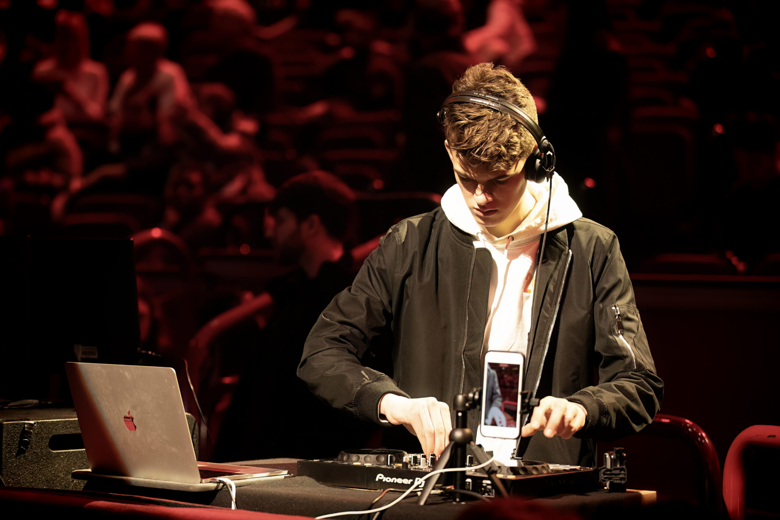 A young DJ wearing headphones performs at a dimly lit venue, using a laptop and DJ controller. A mobile phone is mounted in front of him, and an audience sits blurred in the background under red lighting.