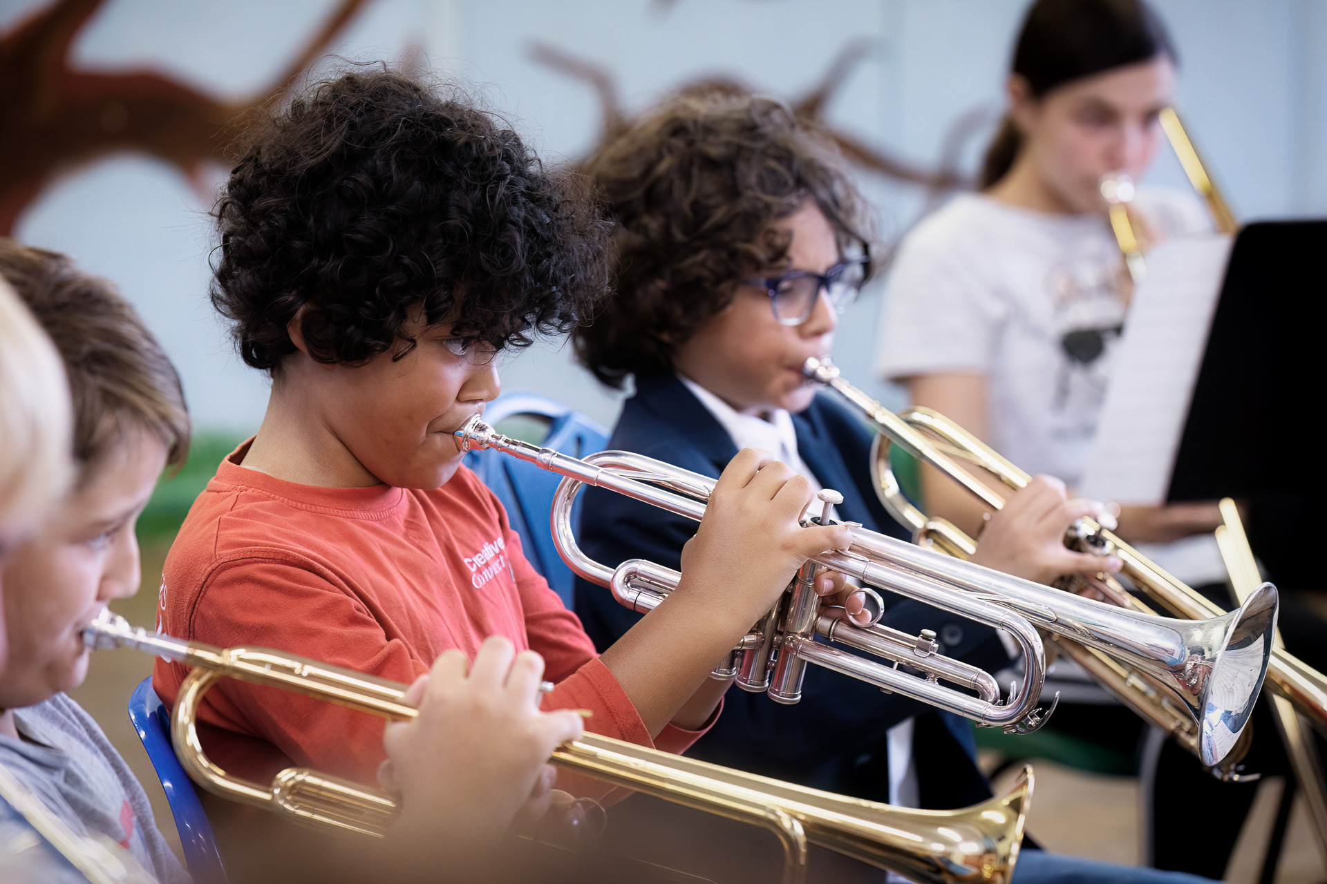Several children sit in a row playing trumpets, focused on their music. The background is blurred, but a sheet of music is visible behind them.