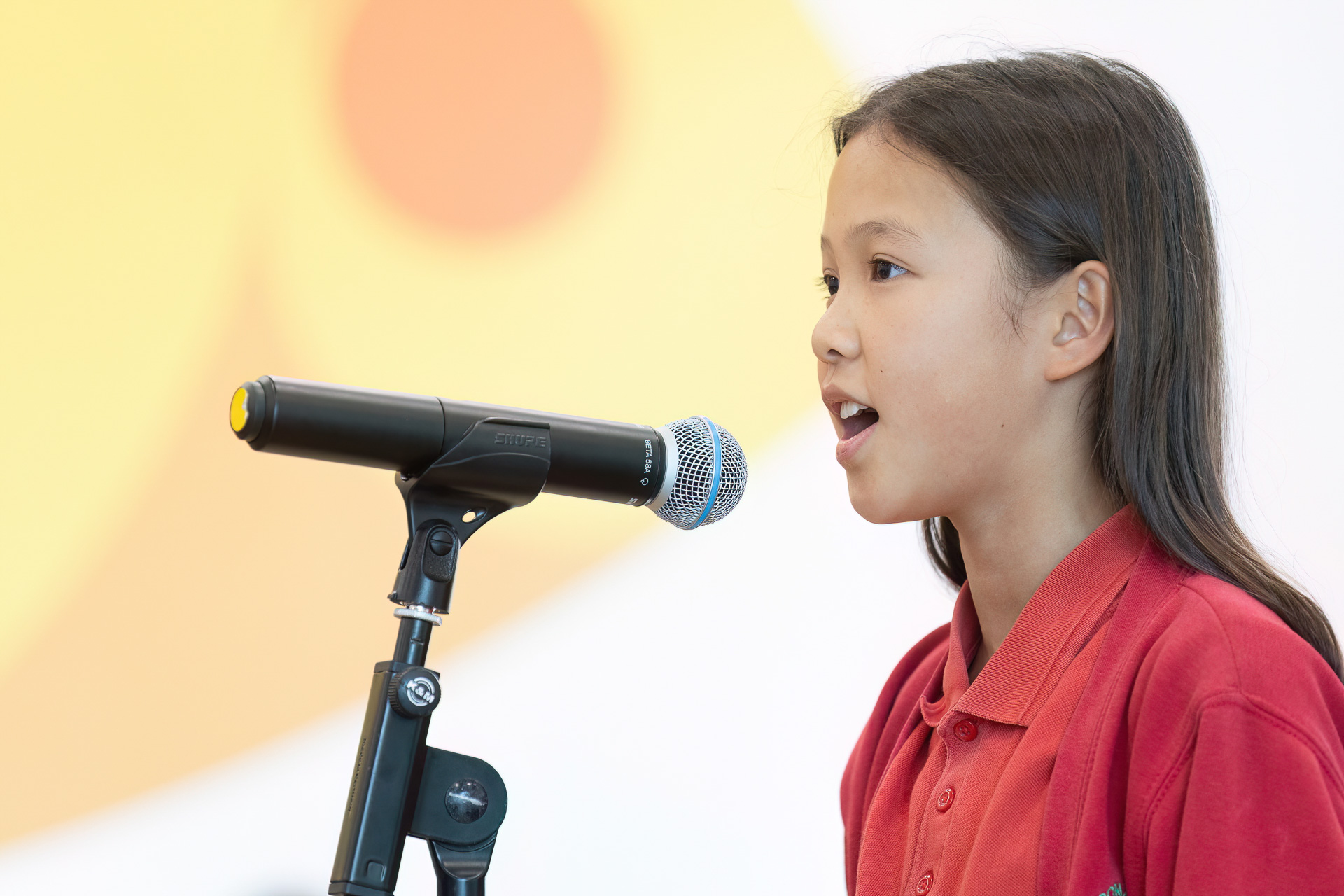 A young girl in a red shirt speaks into a microphone, ready to amplify her voice on stage, with a blurred yellow and orange background behind her.