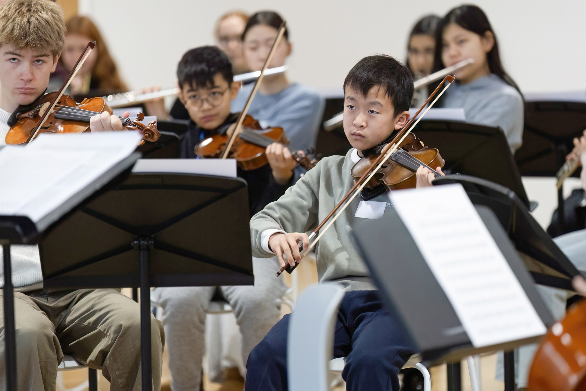 MMF.WIMF.PhilharmoniaWorkshop.01.web A group of young students play violins and other string instruments in an orchestra rehearsal, focussing on their sheet music.