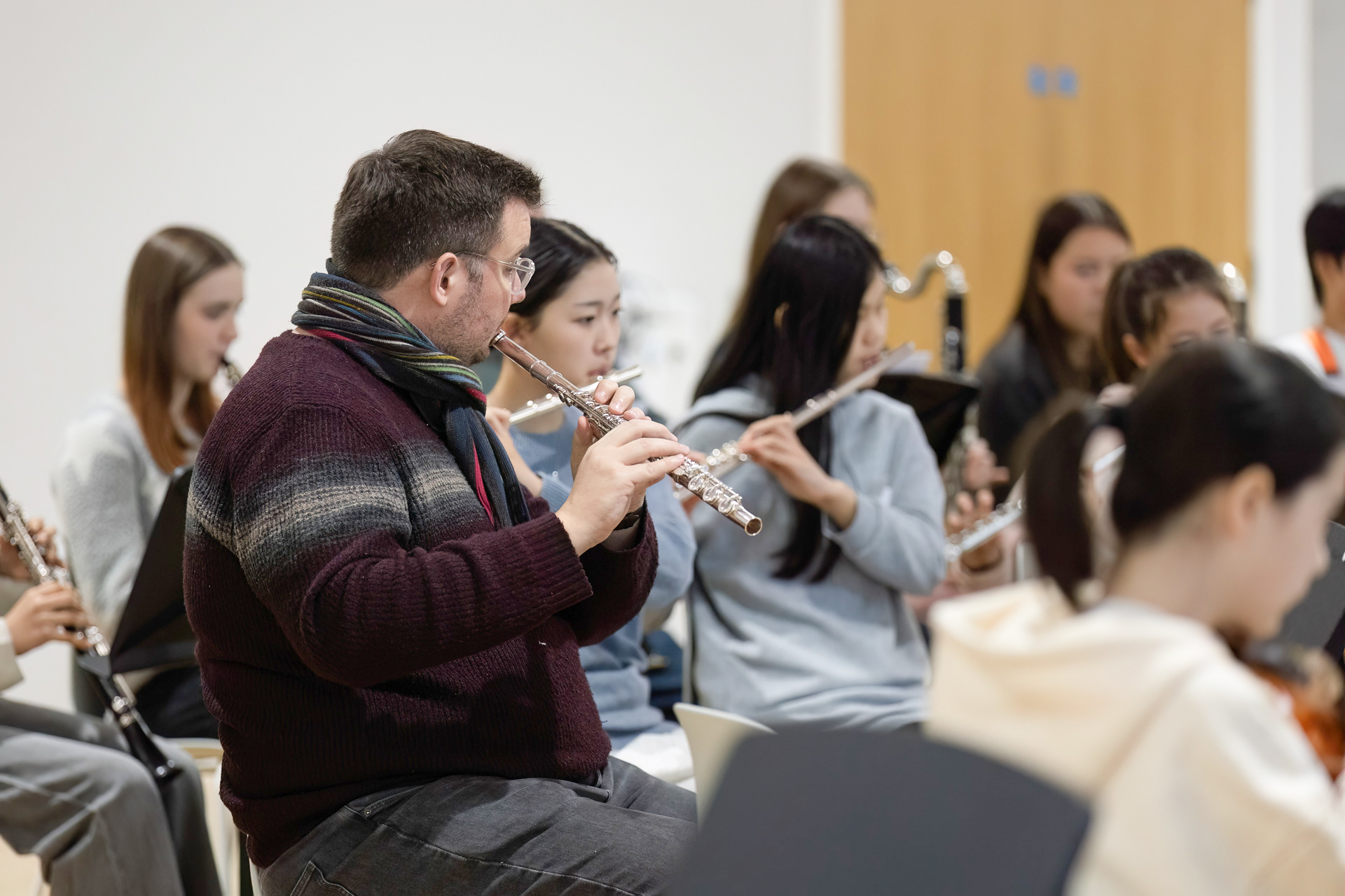 A group of musicians sitting and playing flutes and other woodwind instruments in a rehearsal room, focused on their sheet music. The background is simple with a closed wooden door.