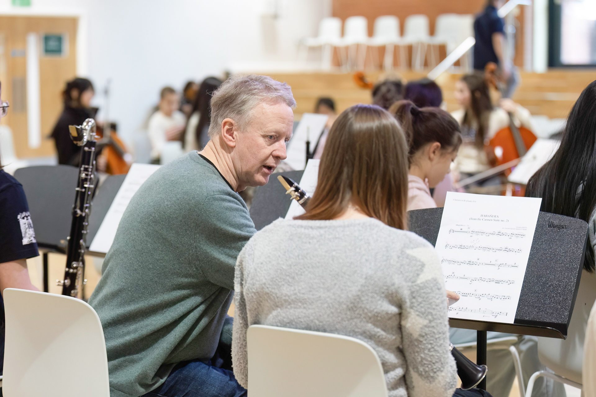 A man and a woman sit with sheet music, facing an orchestra of musicians playing various instruments in a bright rehearsal room. The man appears to be instructing or discussing the music with the woman.
