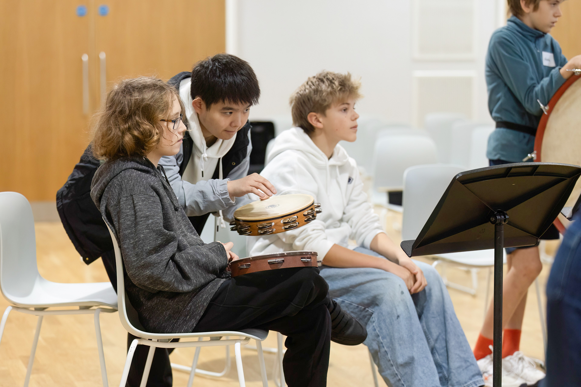 Three boys sit indoors on chairs; two hold tambourines while the third observes. Another pupil stands nearby with a large drum. They appear to be participating in a music lesson or rehearsal. A music stand is in the foreground.