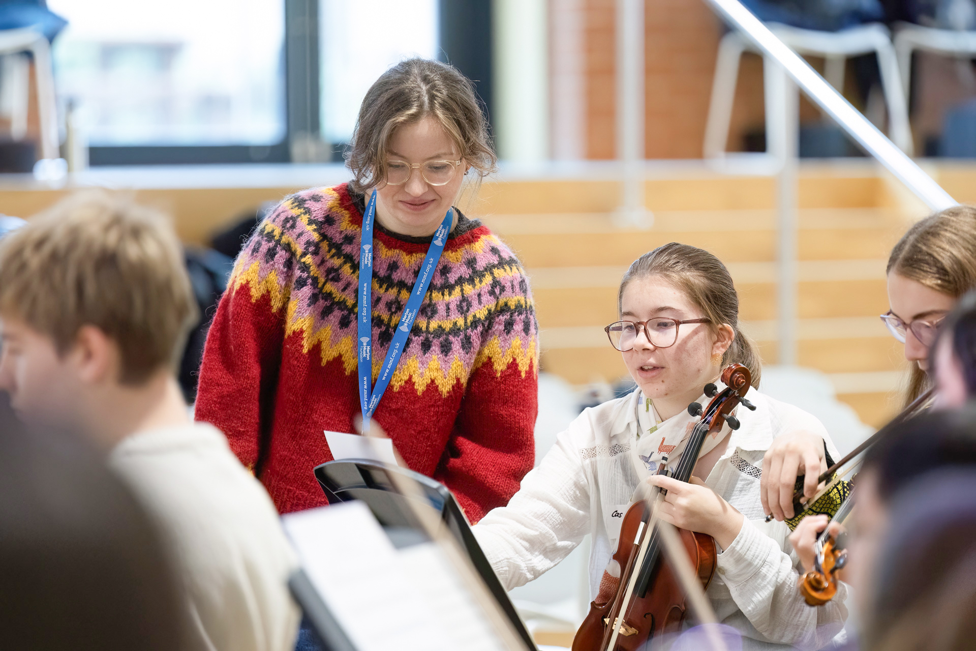 A woman in a red patterned jumper smiles whilst interacting with a girl holding a violin. Other people with instruments are nearby, suggesting a group music practice or lesson indoors.