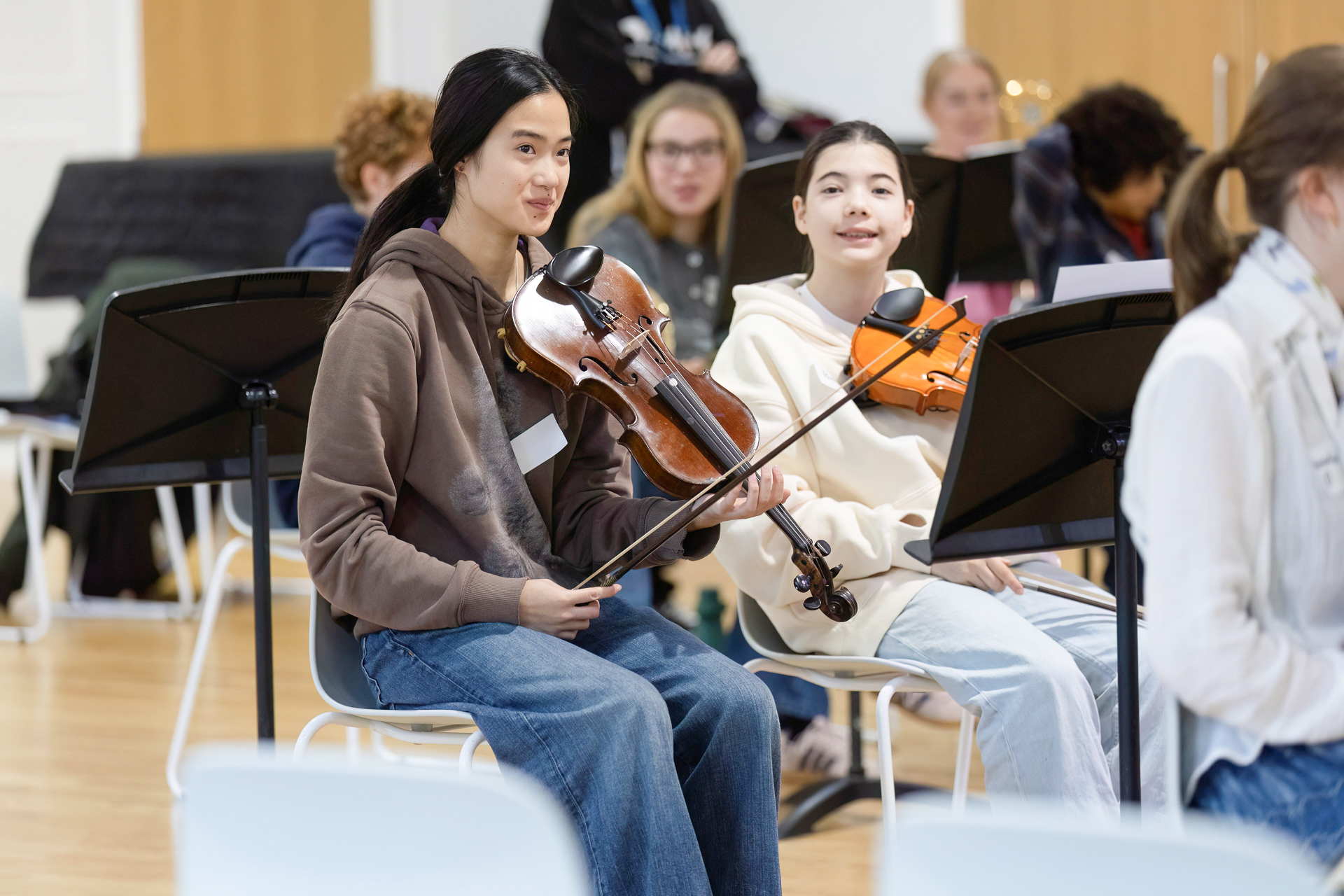 Two young people sit side by side holding violins and smiling in a music classroom. Other students and music stands are visible in the background, suggesting a group rehearsal or lesson.