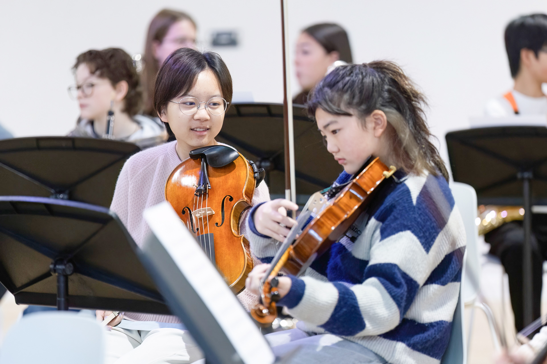 Two young people sitting with violins in an orchestra setting, one looking at the other and smiling whilst the other focuses on her instrument; music stands and other musicians are visible in the background.