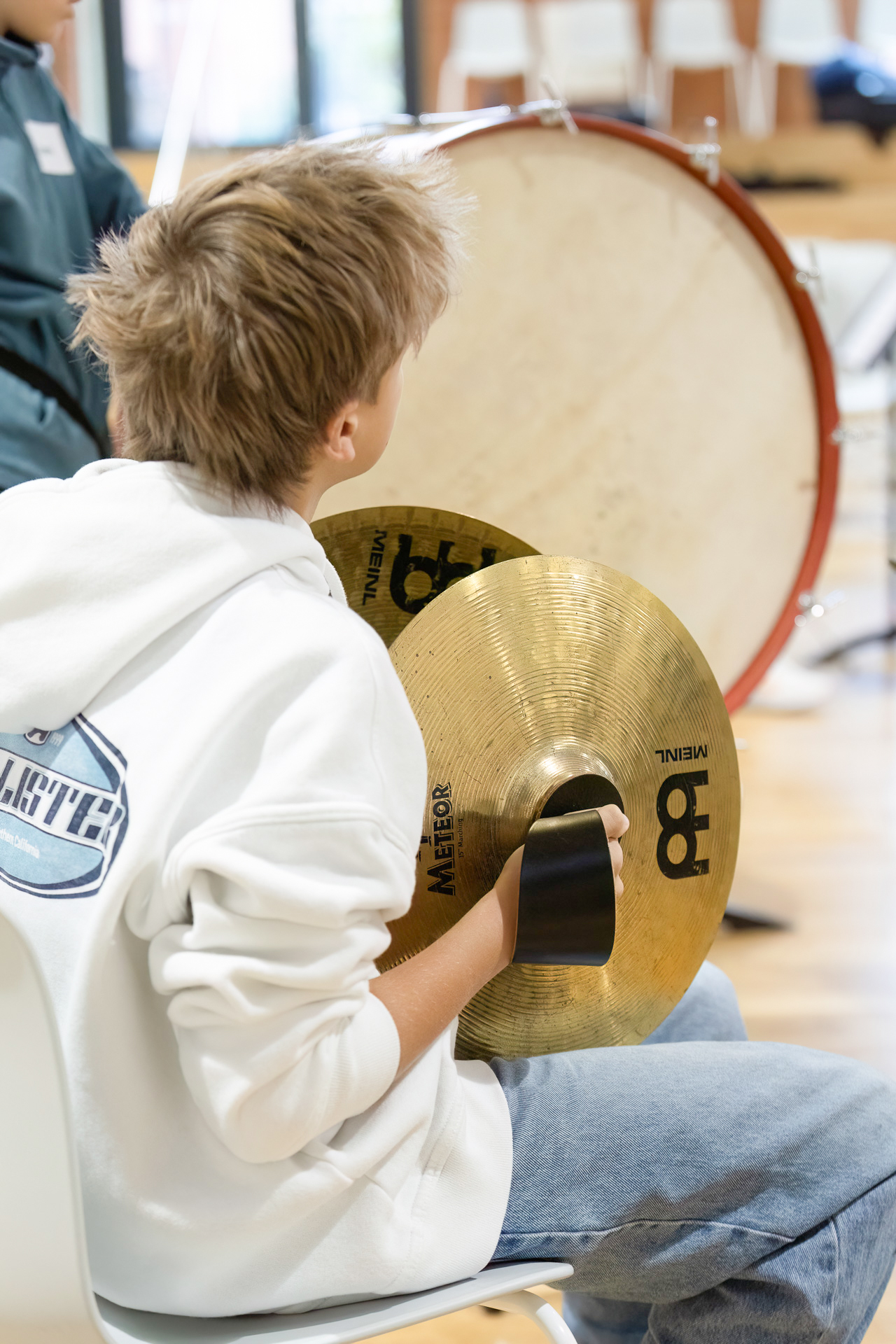 A young person with light brown hair, wearing a white hoodie and jeans, sits on a chair and holds two brass cymbals in their hands. A large bass drum is visible in the background.