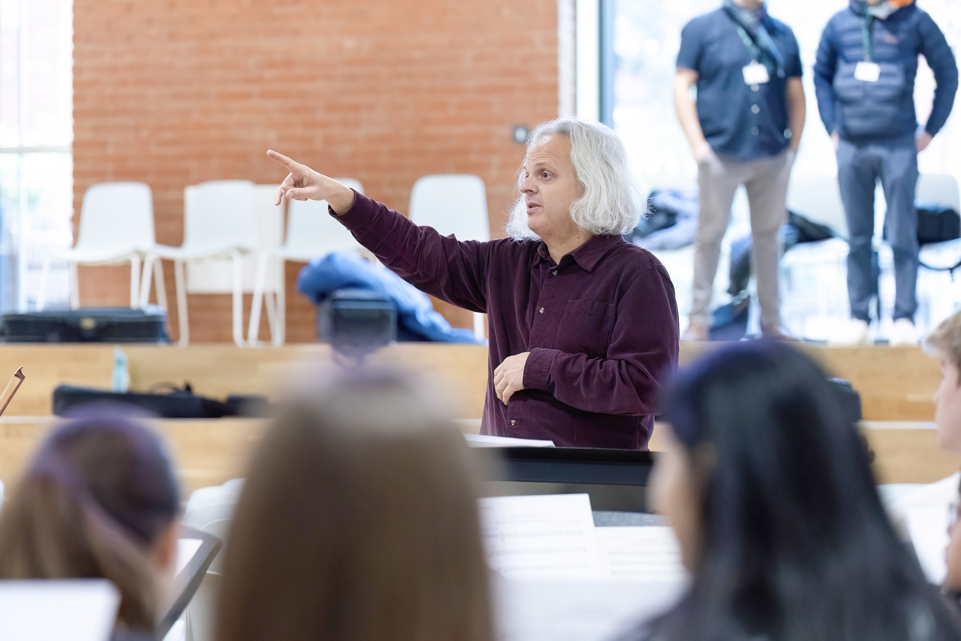 A person with grey hair and a dark shirt stands in front of a group indoors, gesturing with one hand raised and pointing. Several people are seated in the foreground, and others stand in the background.