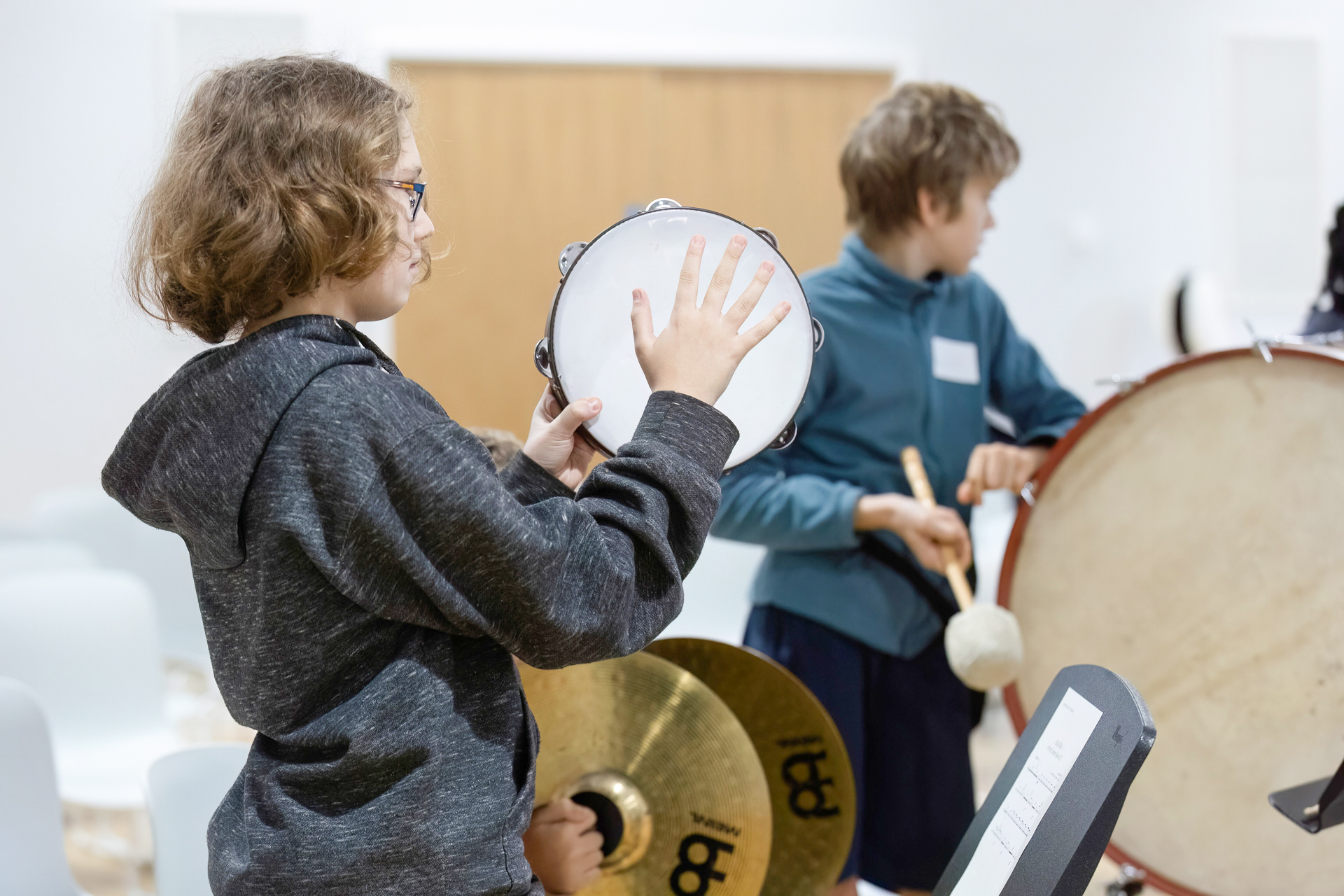 A child wearing glasses plays a tambourine whilst another child in the background plays a bass drum with a beater in a bright classroom or music room.