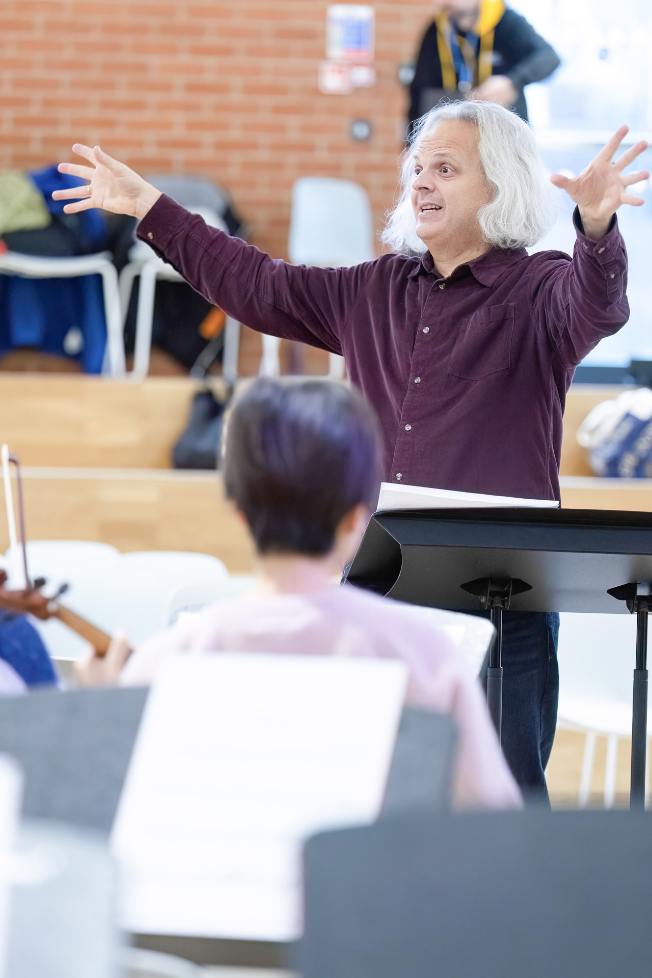 A man with long grey hair enthusiastically conducts musicians in a rehearsal room, raising his arms expressively. A music stand and the back of a musician's head are visible in the foreground.