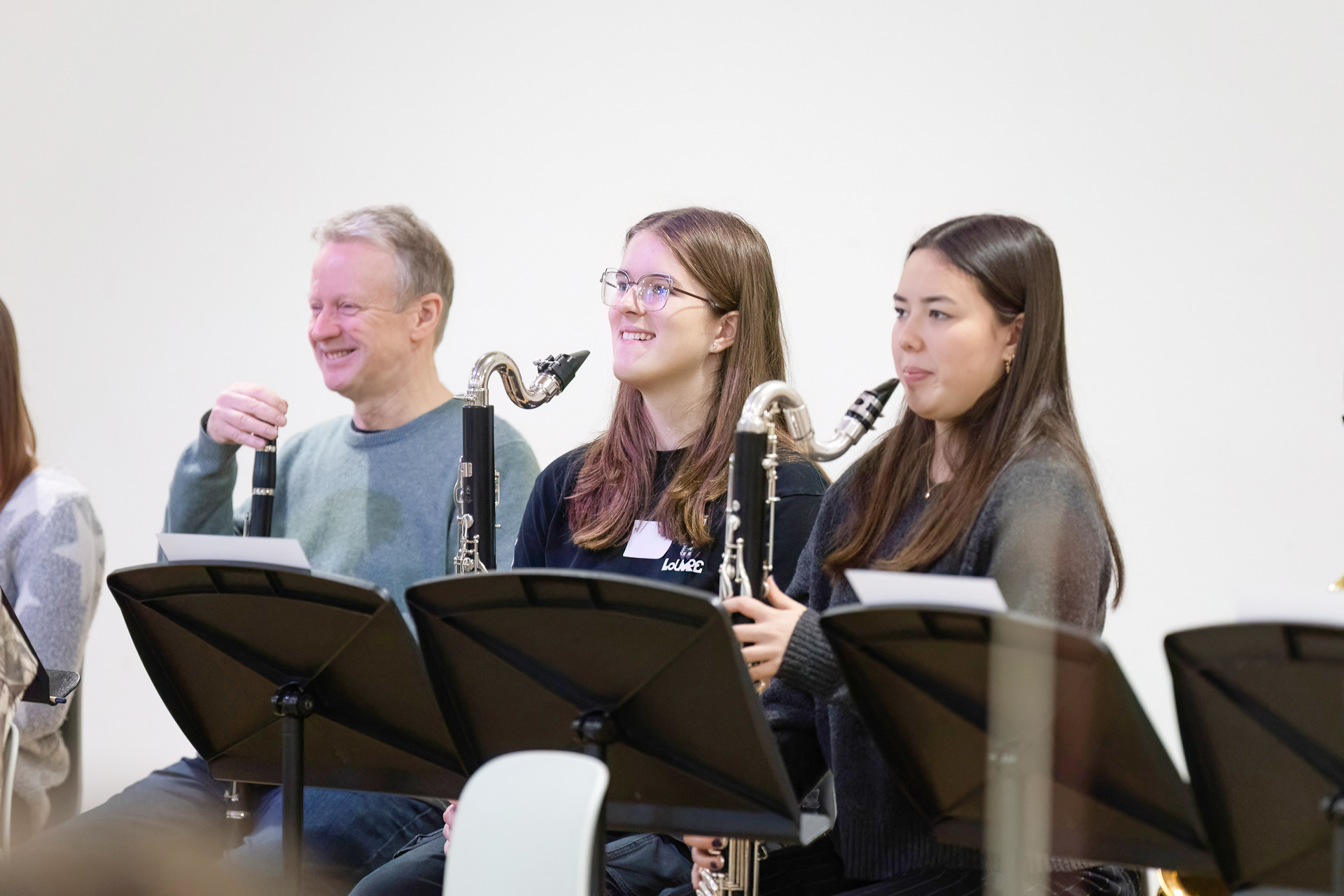 Three people sit behind music stands, holding bass clarinets. They appear to be part of a band or orchestra, smiling and looking ahead. The background is plain and white, keeping the focus on the musicians.