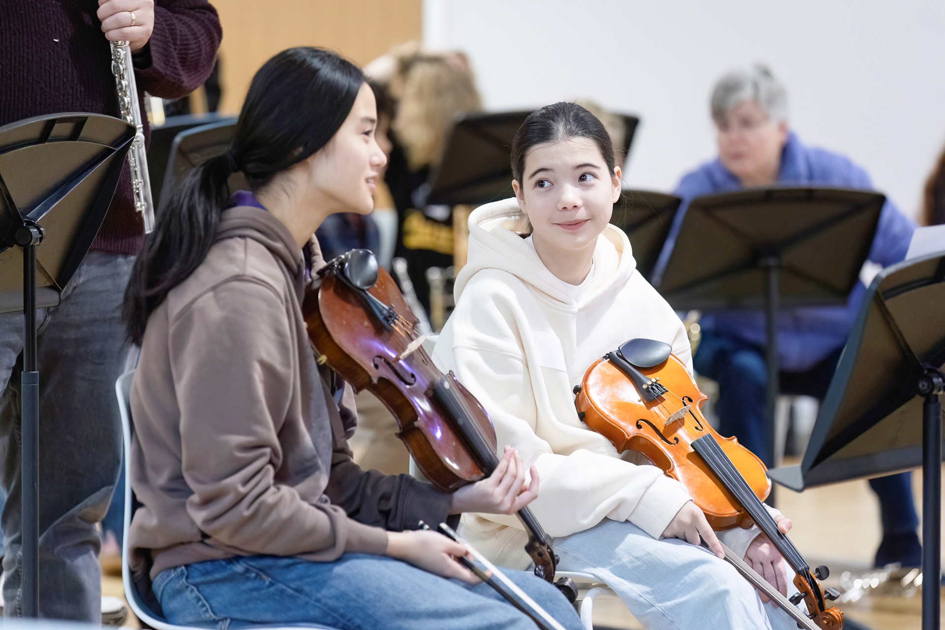 Two young women sit holding violins and smile at each other during a music rehearsal. Other musicians and music stands are visible in the background.
