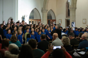 A children's choir dressed in blue shirts sings in a church, led by a conductor, while an audience sits and watches. The church has arched doorways, tall columns, and soft lighting.