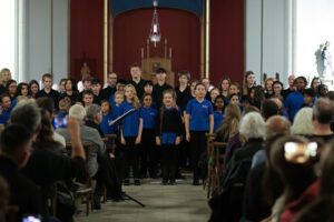A children's choir, mostly in blue shirts, sings in a church whilst an audience listens and takes photos. The choir stands in rows facing the seated crowd, with candles and a cross behind them on the altar.