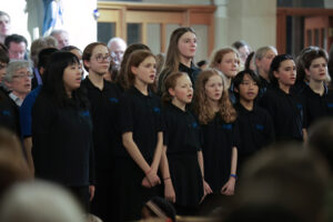 A group of children and adults in matching black shirts sing together in a choir inside a well-lit building, possibly a church or concert hall, with a blurred audience in the foreground.