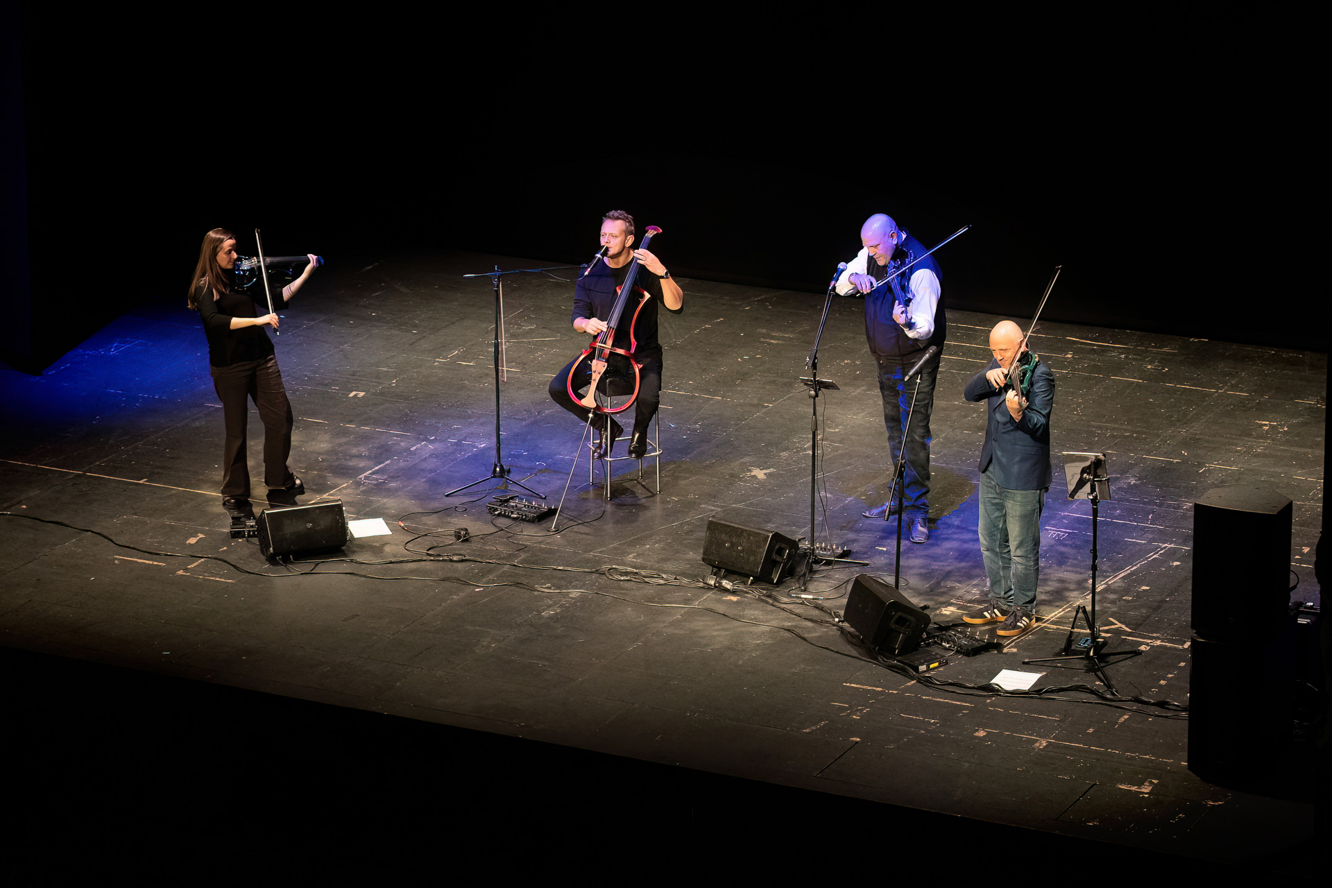Four musicians perform on a dimly lit stage; three play violins and one plays a cello. Microphones and music stands are set up in front of them, and cables are visible on the floor.