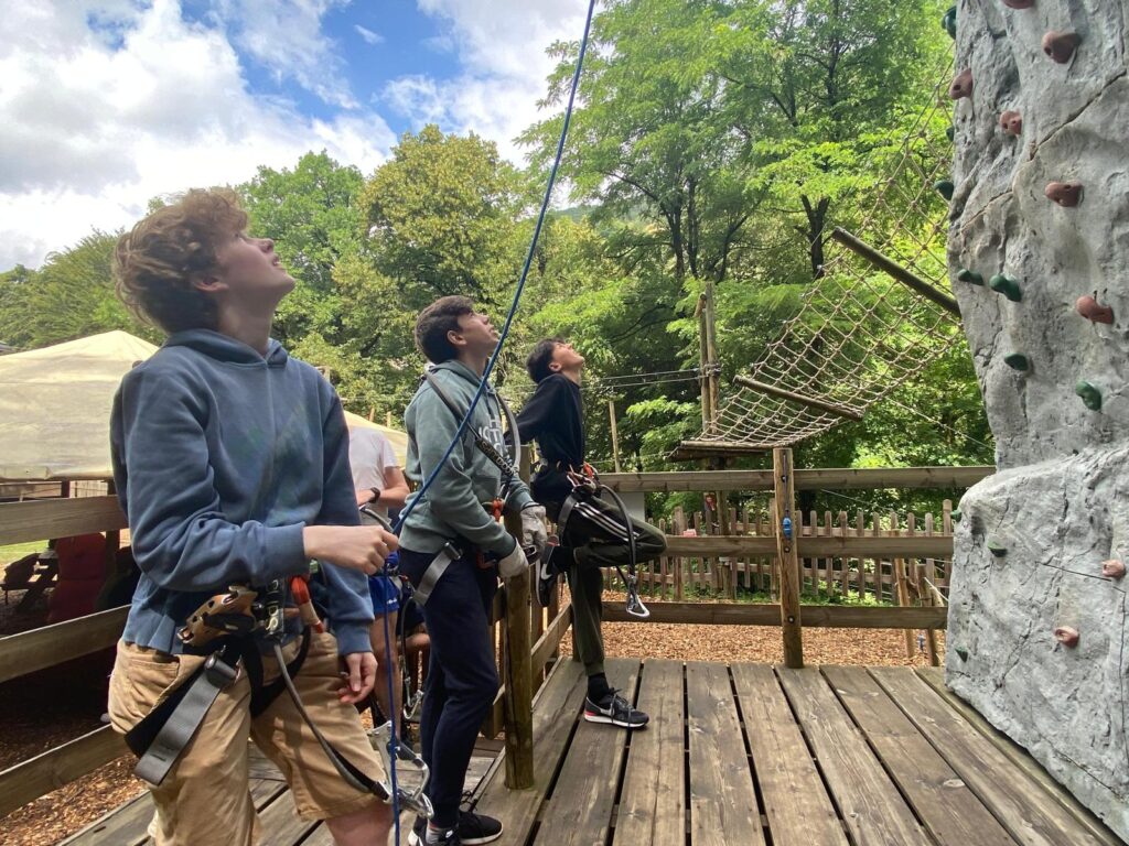 Three teenagers wearing harnesses stand on a wooden platform, preparing for rock climbing during the Moselle Valley Tour 2026. Trees and ropes course elements fill the background of this outdoor adventure scene.