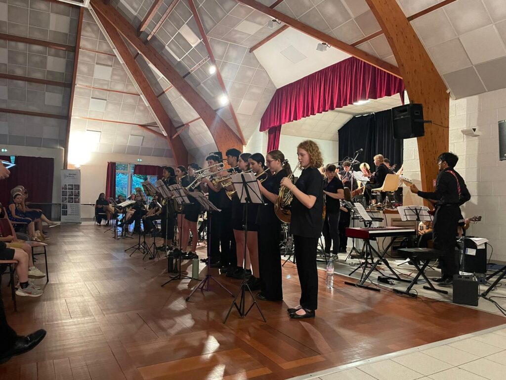 A large student jazz band performs on stage in a wooden-beamed hall during the Moselle Valley Tour 2026, with audience members seated on the left. Musicians play brass, wind, and rhythm instruments, using sheet music stands arranged in front of them.