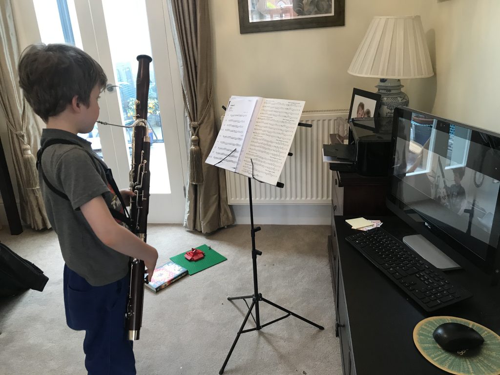 A young boy stands in a South Wimbledon living room, playing a bassoon while looking at sheet music on a stand. A computer with a video call open is on a desk nearby, suggesting an online music lesson.