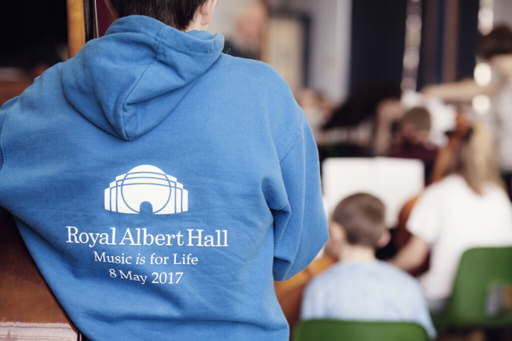 A person in a blue hoodie with the Royal Albert Hall logo and Music is for Life, 8 May 2017 printed on the back, standing among children in a classroom or rehearsal setting.