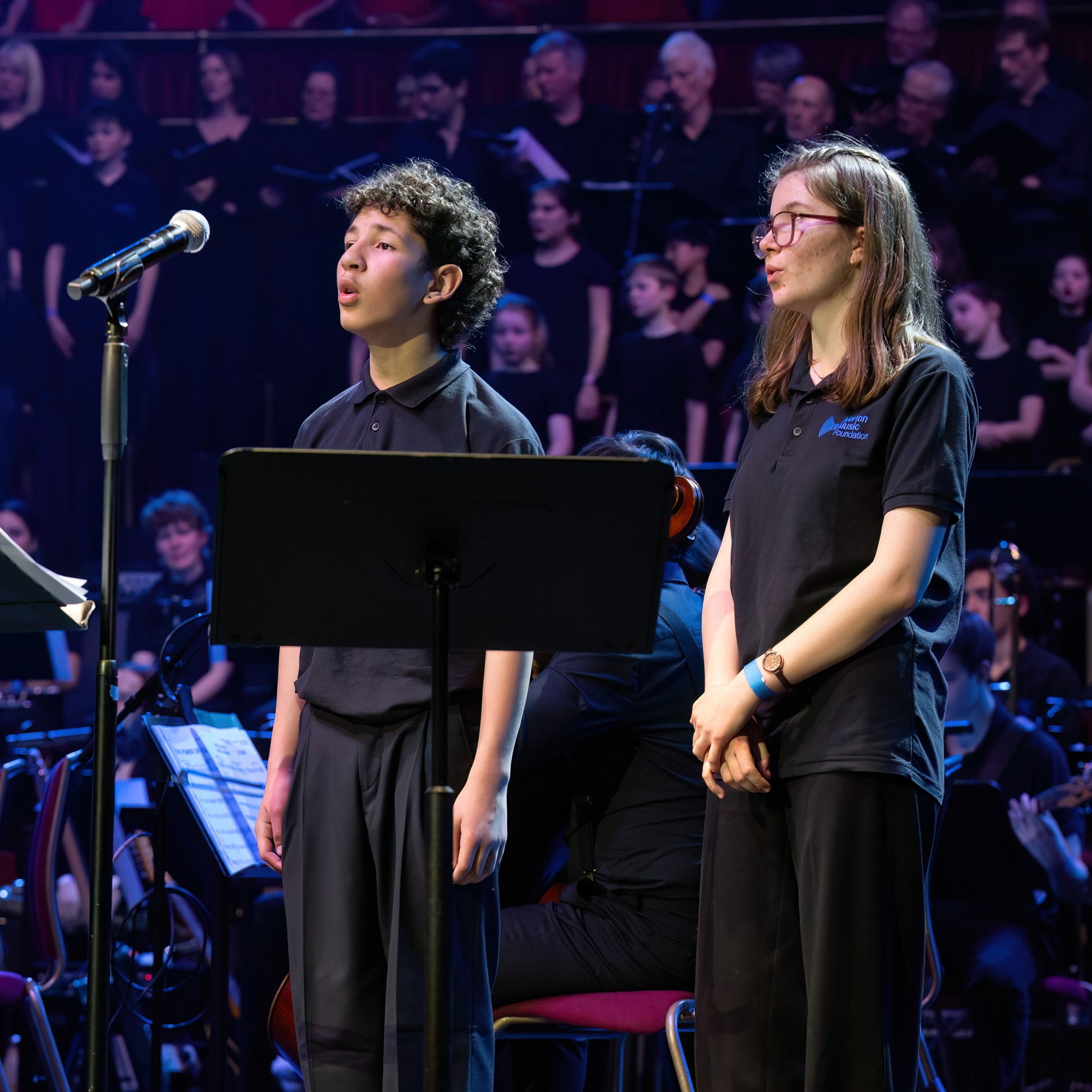 Two young people in black outfits stand on stage singing into a microphone, with sheet music on a stand before them. The Senior Guitar Ensemble, along with the orchestra and choir, is visible in the background under stage lighting.