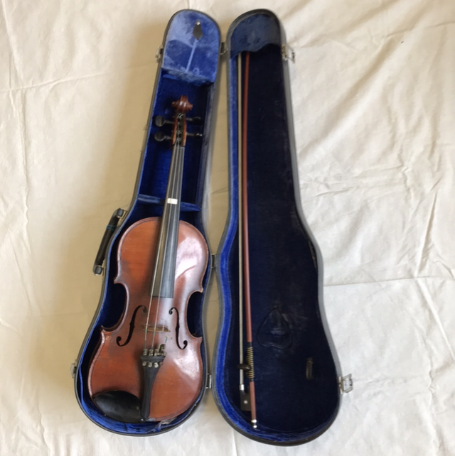 A wooden violin rests in a blue velvet-lined hard case, beside a bow in its own compartment. The open case, displayed on white fabric, represents the quality instruments supported by the Merton Music Foundation.