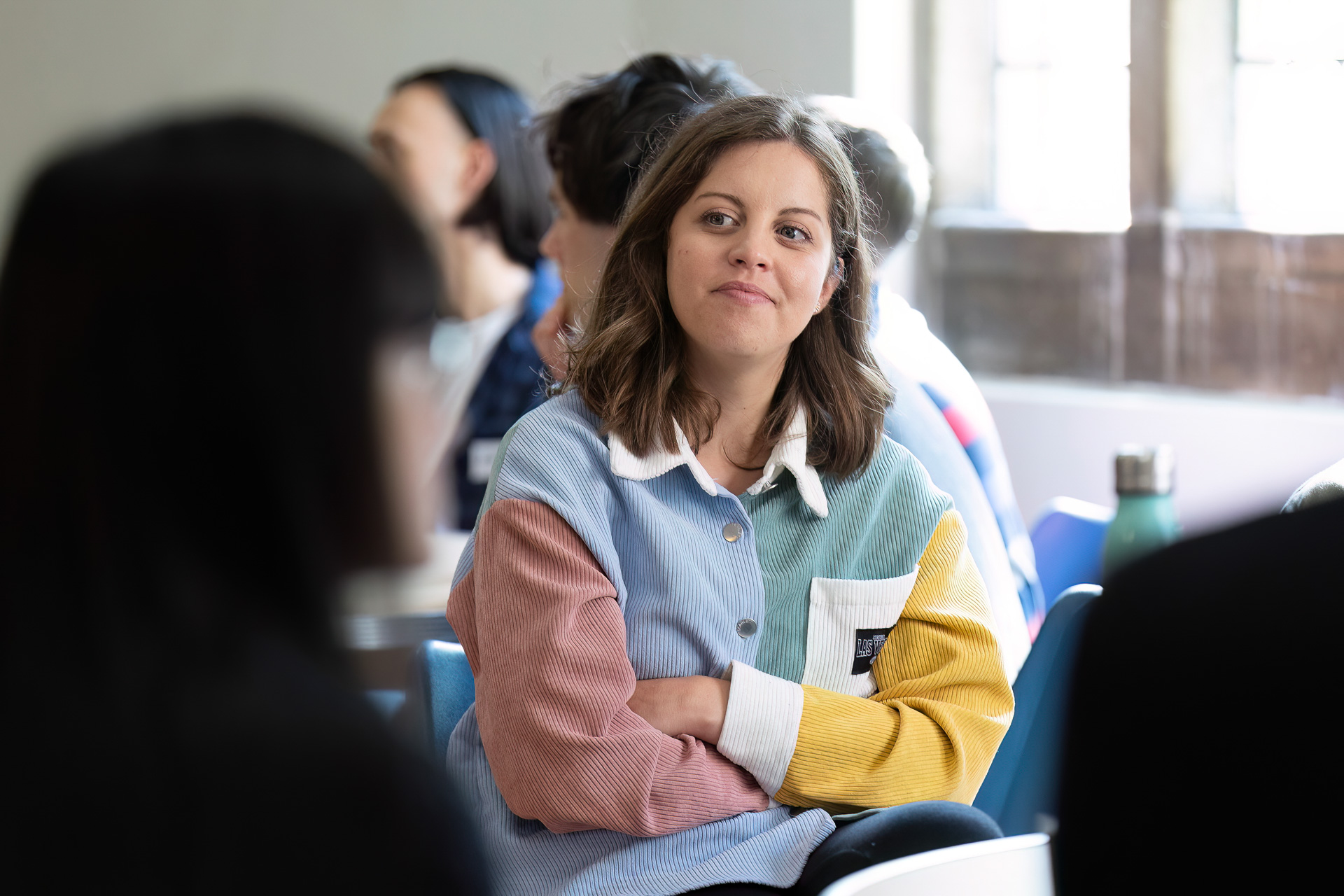 A woman with shoulder-length brown hair, wearing a colourful, patchwork shirt, sits with her arms crossed and smiles slightly whilst listening to someone in a bright room with other people blurred in the background.