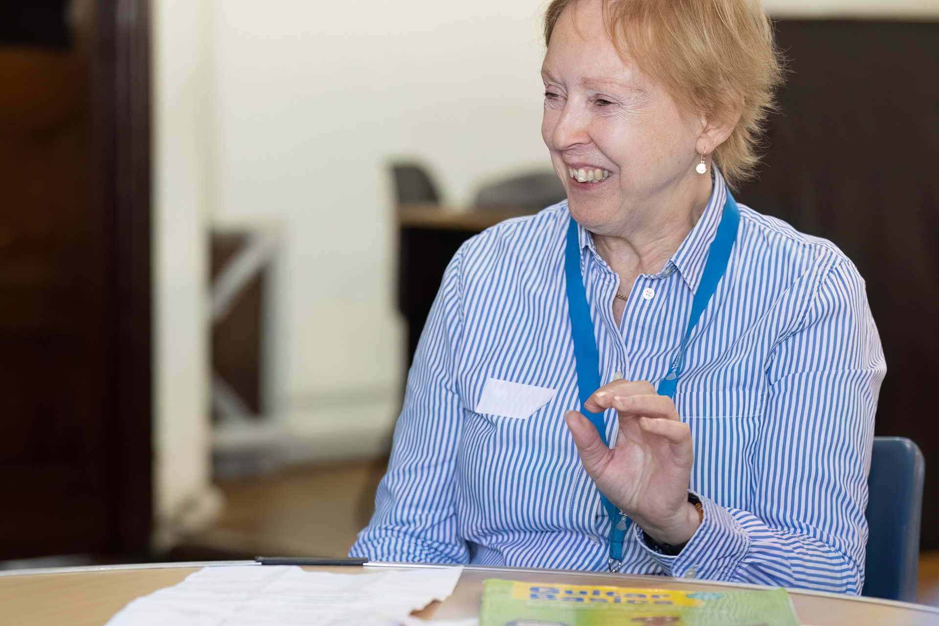 An older woman wearing a blue lanyard and a striped shirt sits at a table, smiling and gesturing with her hand. Papers and a book are on the table in front of her. The background is out of focus.