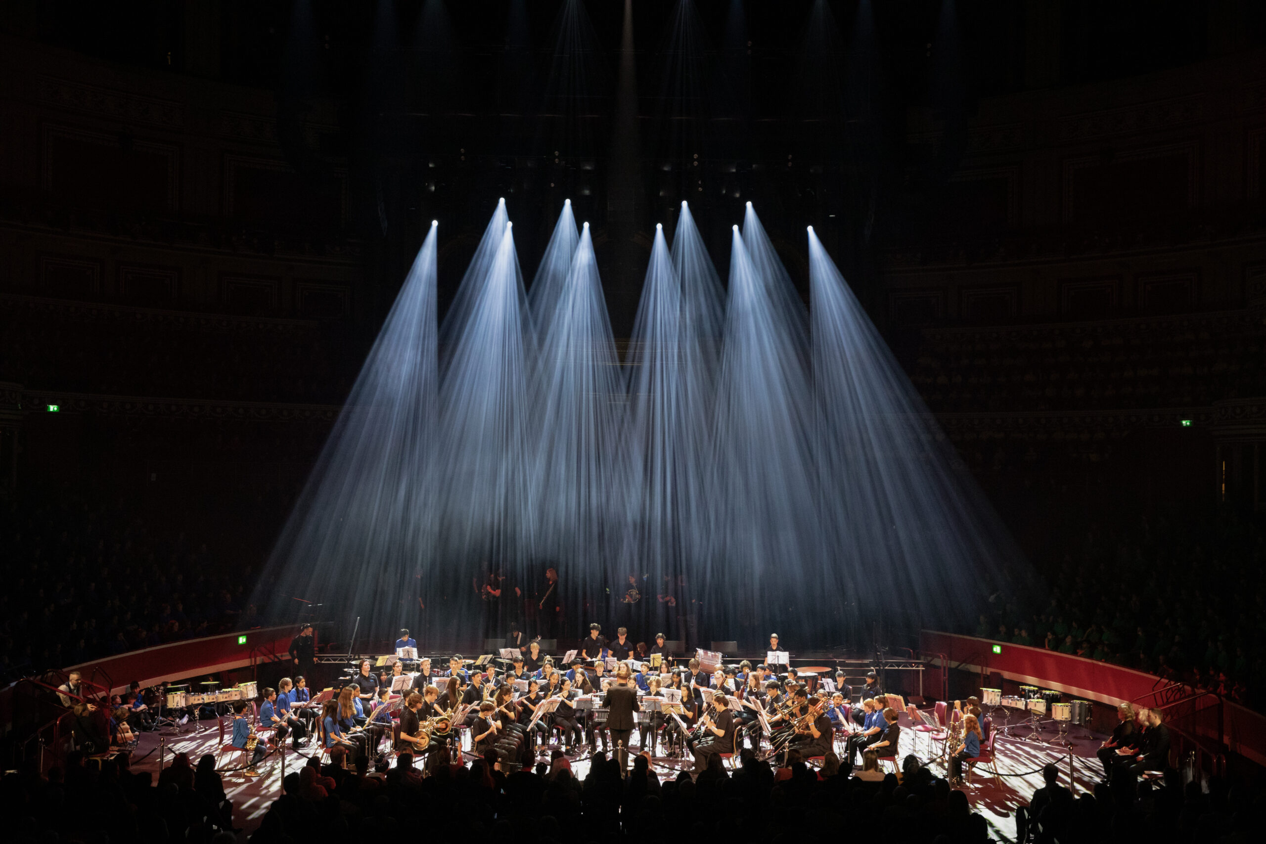 An orchestra performs on a large stage under dramatic, bright white spotlights that shine down in beams, illuminating the musicians whilst the audience sits in darkness.