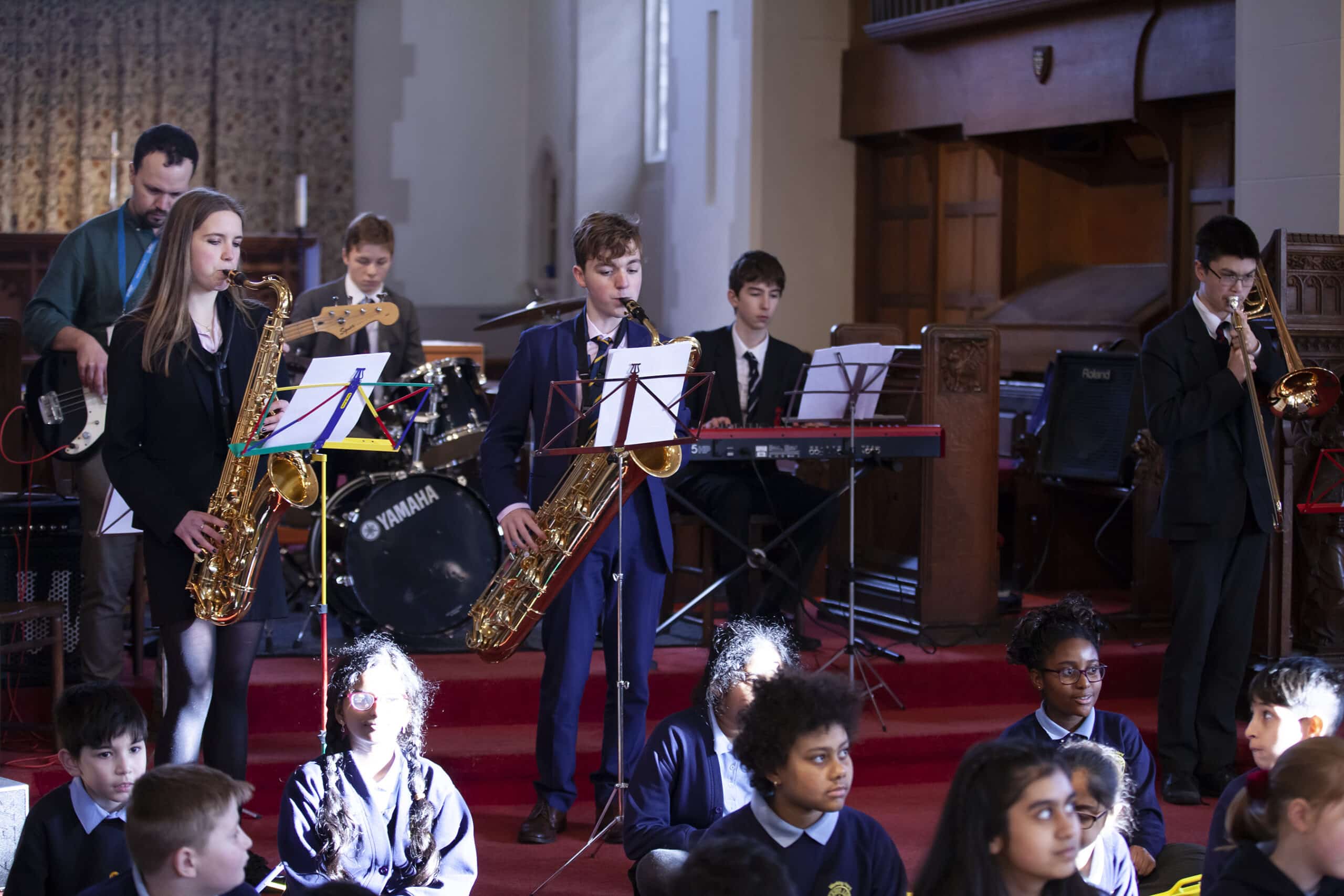 A school band performs on stage with students playing saxophones, trombone, guitar, keyboard, and drums, whilst other students sit on the floor in front, watching attentively. The setting appears to be a church or hall.