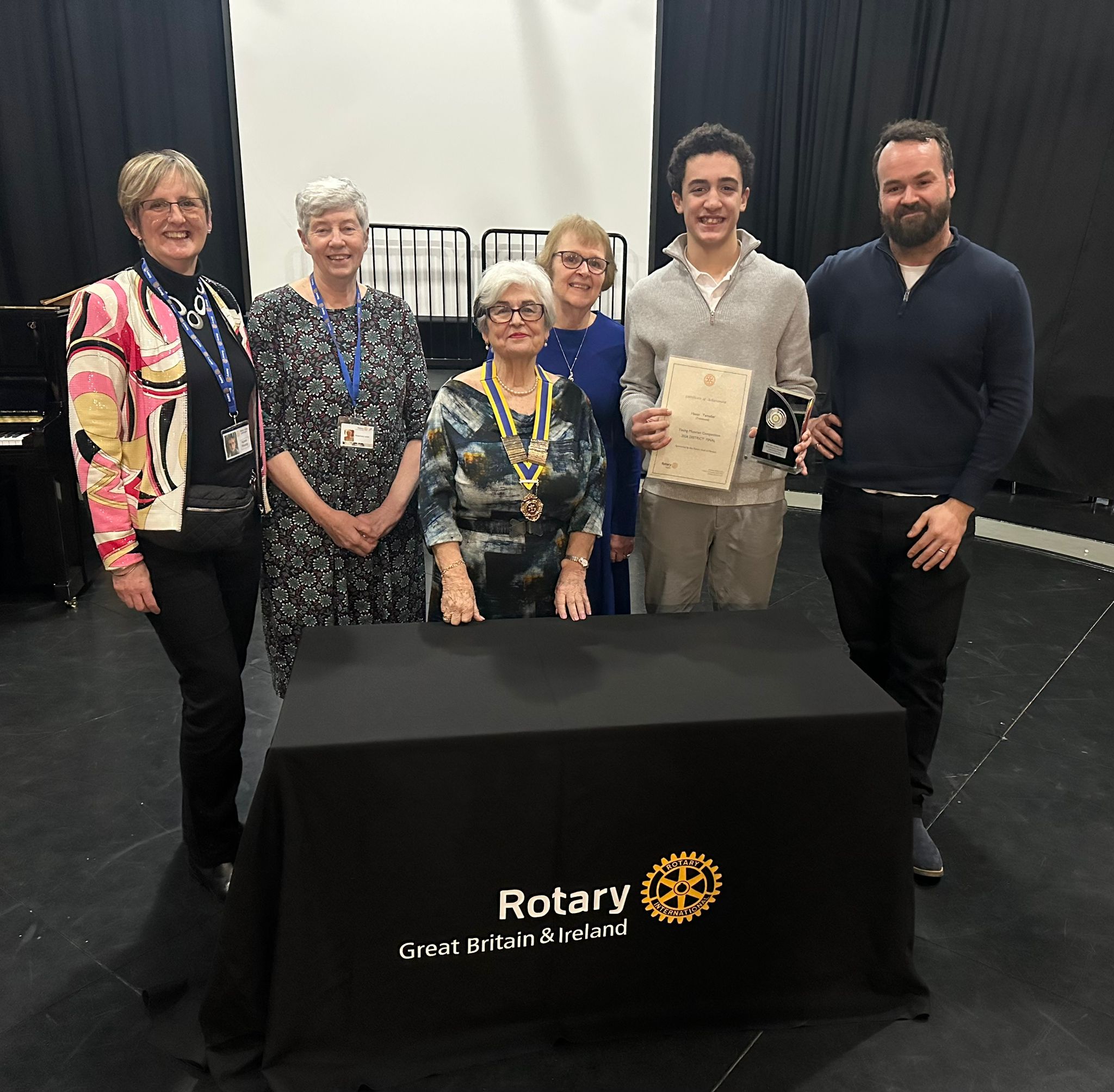 Six people stand behind a table with a “Rotary Great Britain & Ireland” logo. A young man, smiling, holds a certificate and medal from the Philharmonia Orchestral Workshop as others pose proudly beside him in a formal indoor setting.