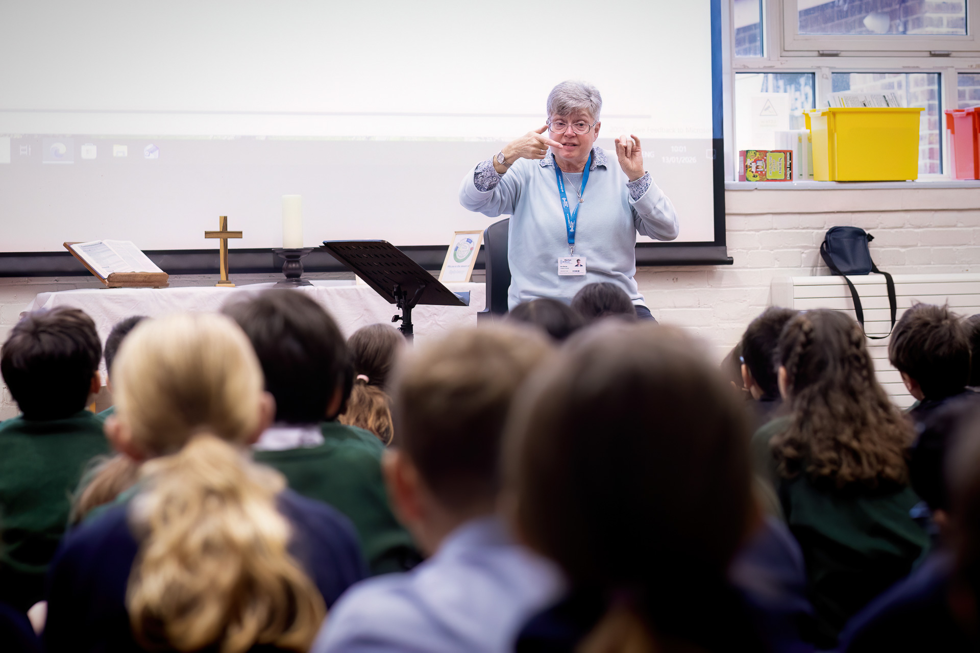 A teacher sits at the front of a large classroom or hall, gesturing as she leads a singing workshop. A cross, candle, and book rest on a table beside her whilst the students sing.