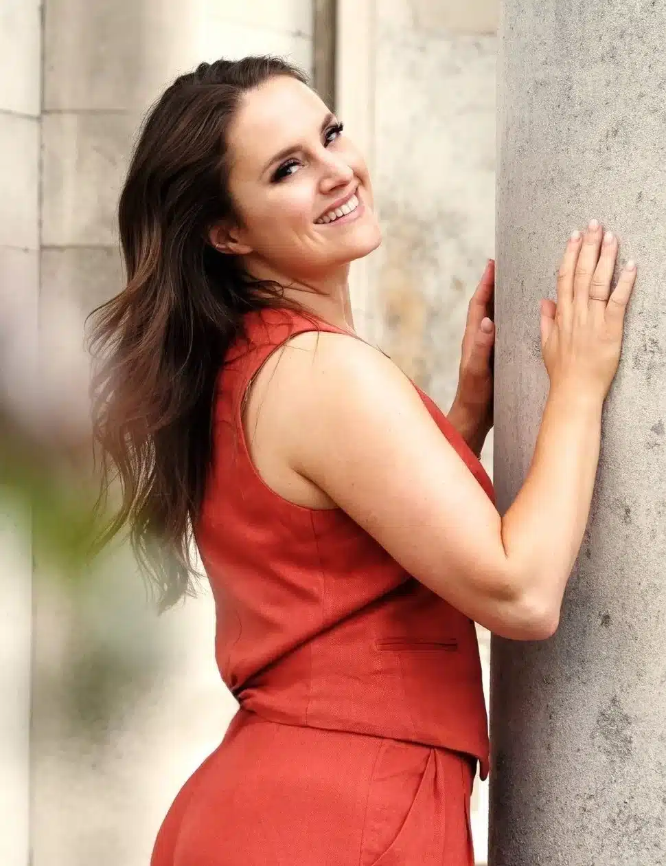 A woman with long brown hair, wearing a sleeveless rust-coloured outfit, smiles whilst leaning against a large stone pillar outdoors, celebrating the success of the Ticket Bursary Scheme.