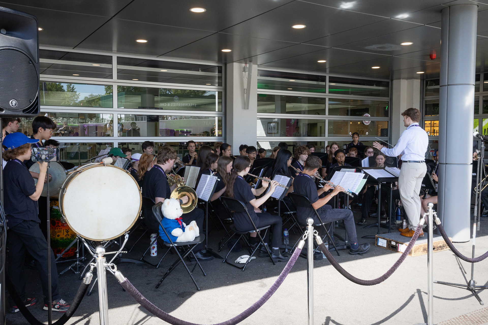 A school band performs outdoors under a covered area, led by a conductor. Musicians play various instruments whilst seated; a large drum and a stuffed bear are visible in the front row. Onlookers and equipment surround the group.