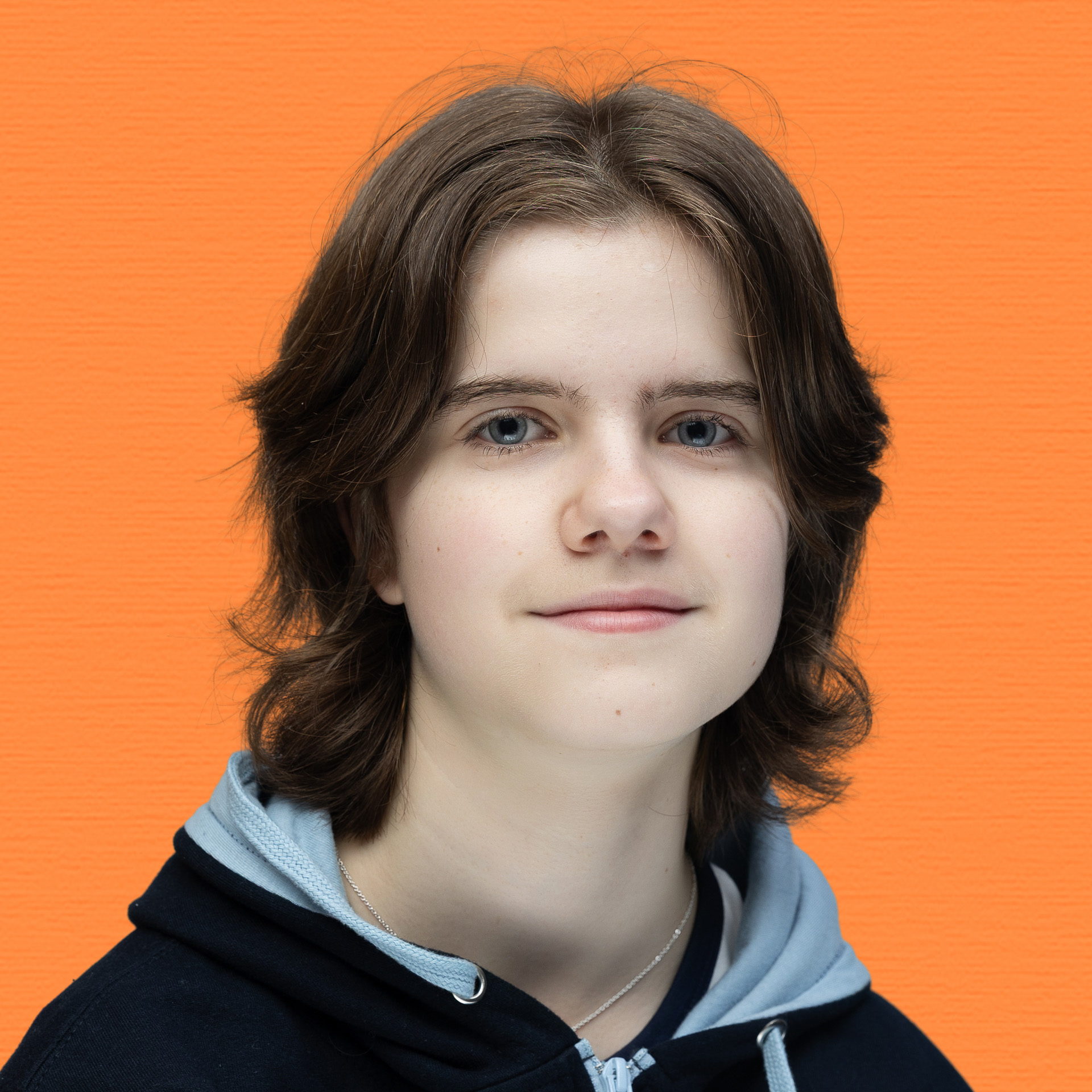 A young person with medium-length brown hair and blue eyes, wearing a black and grey hoodie, smiles gently in front of an orange textured background, proudly representing the Youth Council.