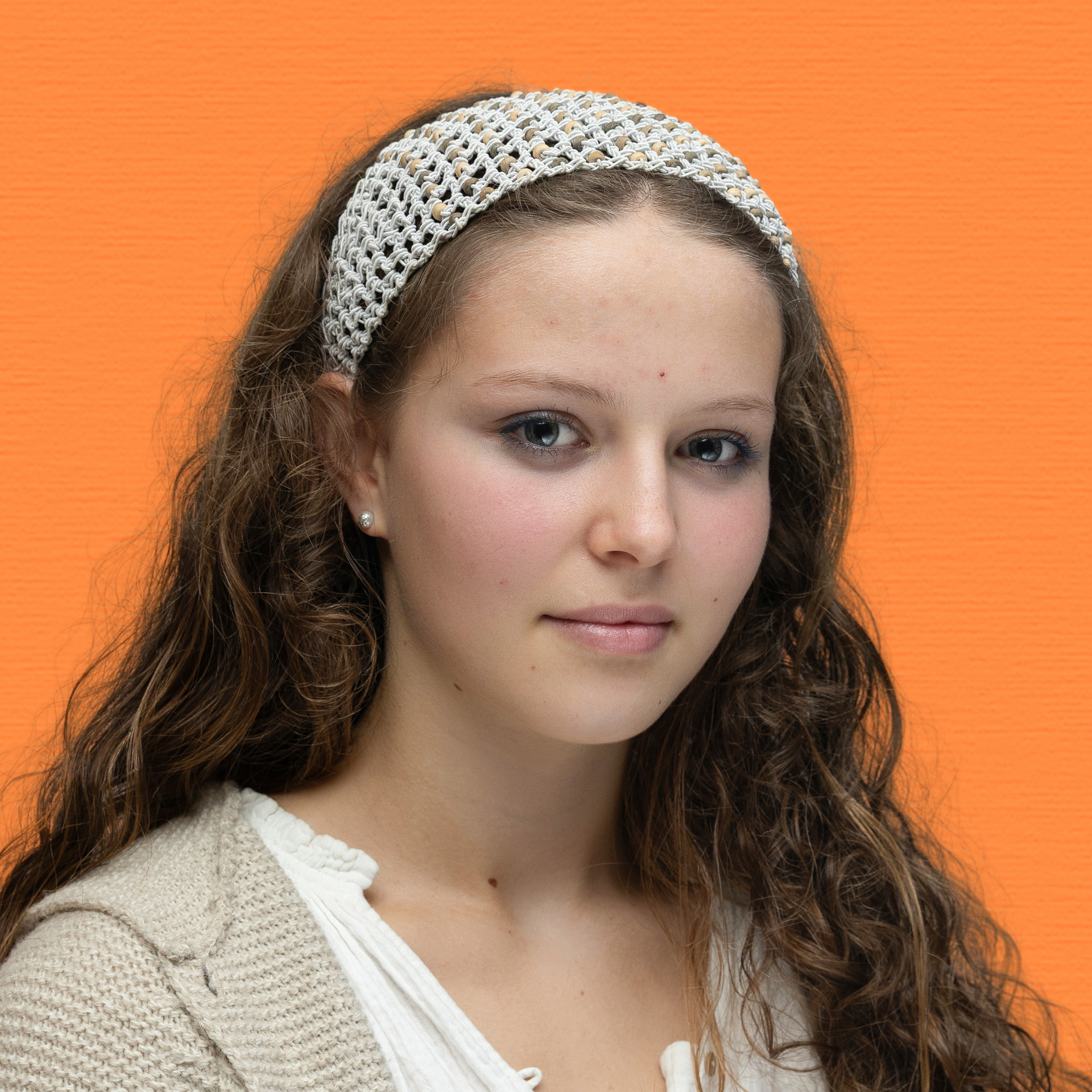 A young woman with long curly hair, wearing a white crocheted headband and a cream-coloured top, poses in front of an orange background. She has a neutral expression and light make-up, representing the poised spirit of the Youth Council.