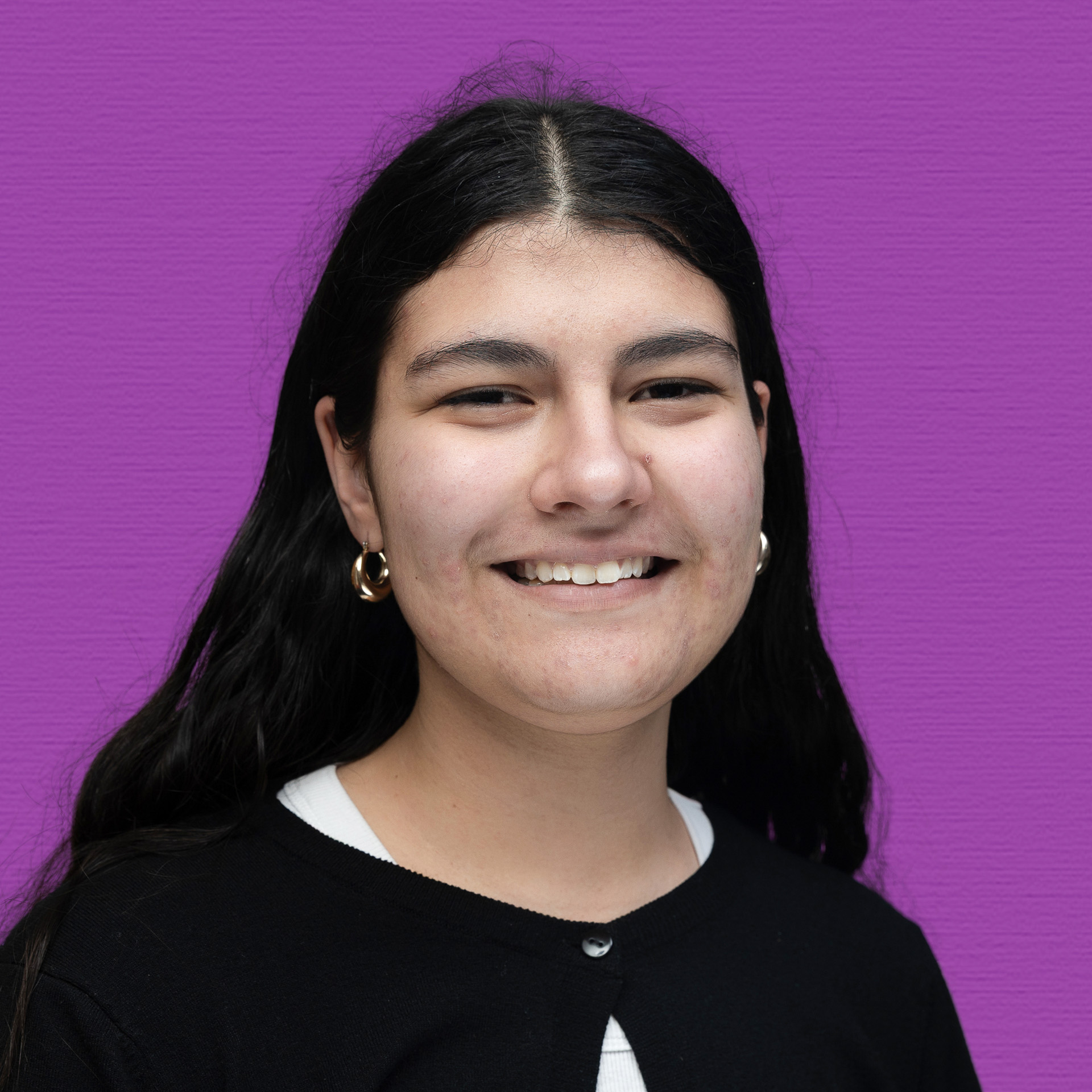 A young woman with long dark hair, wearing gold hoop earrings, a black cardigan, and a white top, smiles in front of a bright purple background—representing the vibrant spirit of the Youth Council.