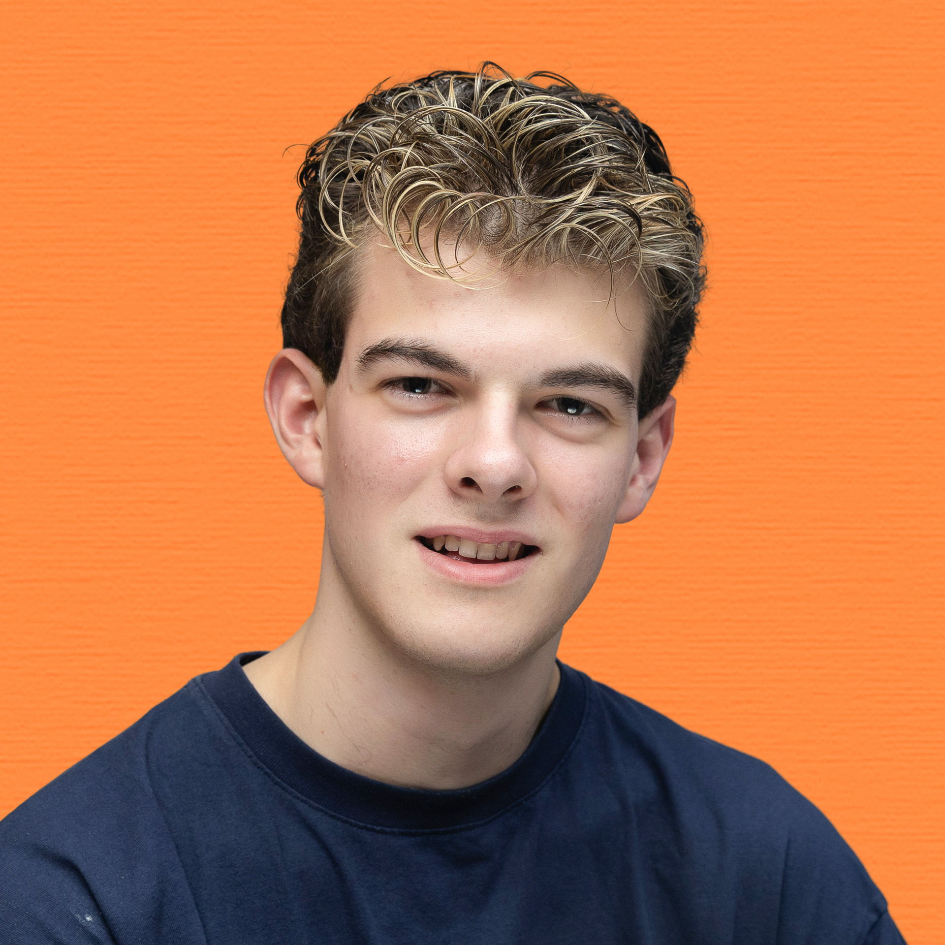 A young man with short, curly, highlighted hair wearing a dark blue T-shirt stands in front of a textured orange background, smiling slightly at the camera—a confident member of the Youth Council.