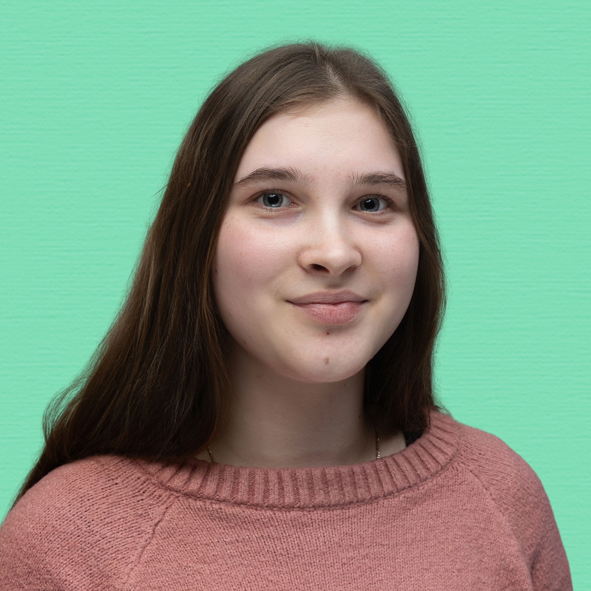 A young woman with long brown hair, wearing a pink jumper, smiles slightly whilst standing in front of a solid mint green background, representing the spirit of the Youth Council.