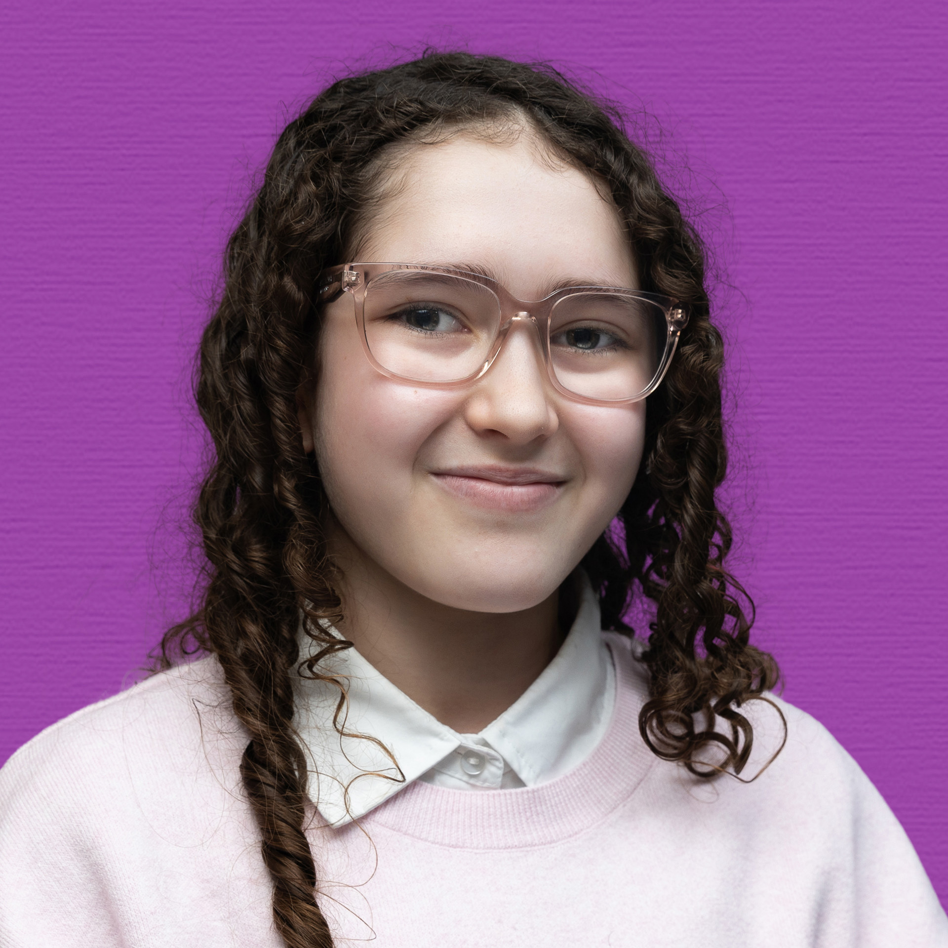 A young person with long curly brown hair and glasses is smiling softly, wearing a light pink jumper over a white collared shirt, standing in front of a purple background, representing the Youth Council.