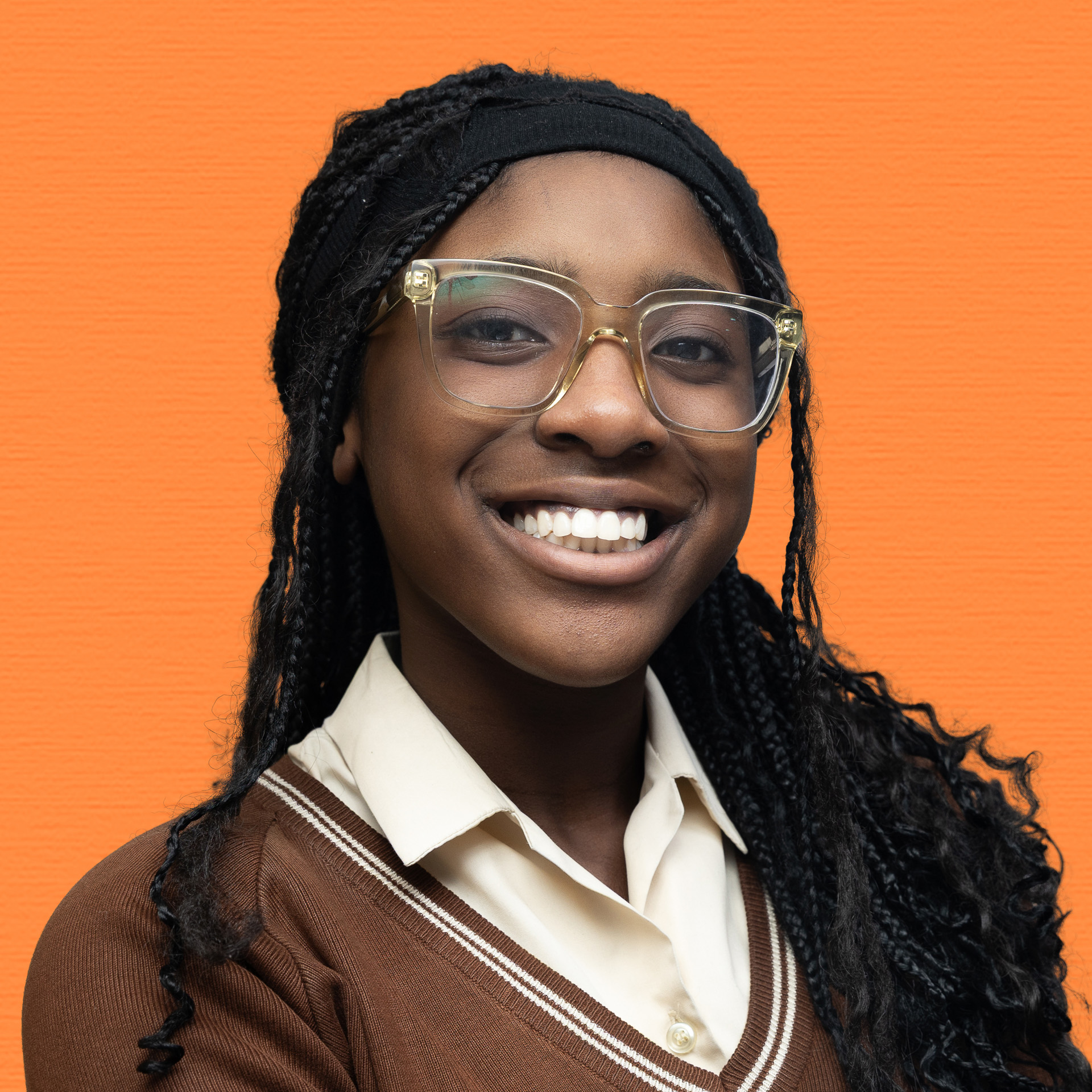A young woman with long plaited hair, wearing large clear glasses and a brown jumper over a collared shirt, smiles in front of an orange background, representing the Youth Council with confidence and optimism.