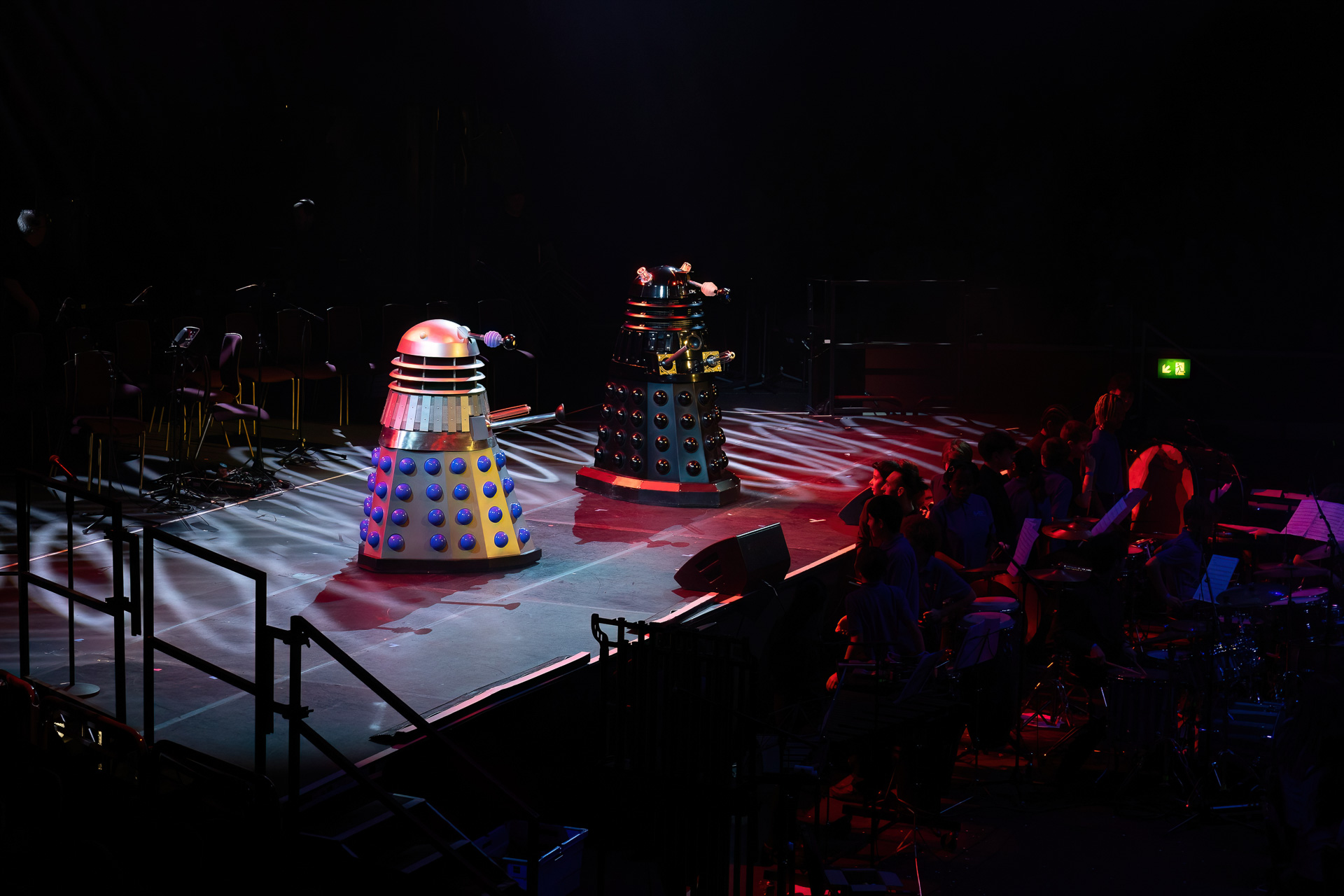 Two Dalek robots from Doctor Who are on a stage at Drumathon 2025, illuminated by colourful lights, with musical instruments and an orchestra to the right and empty chairs to the left.
