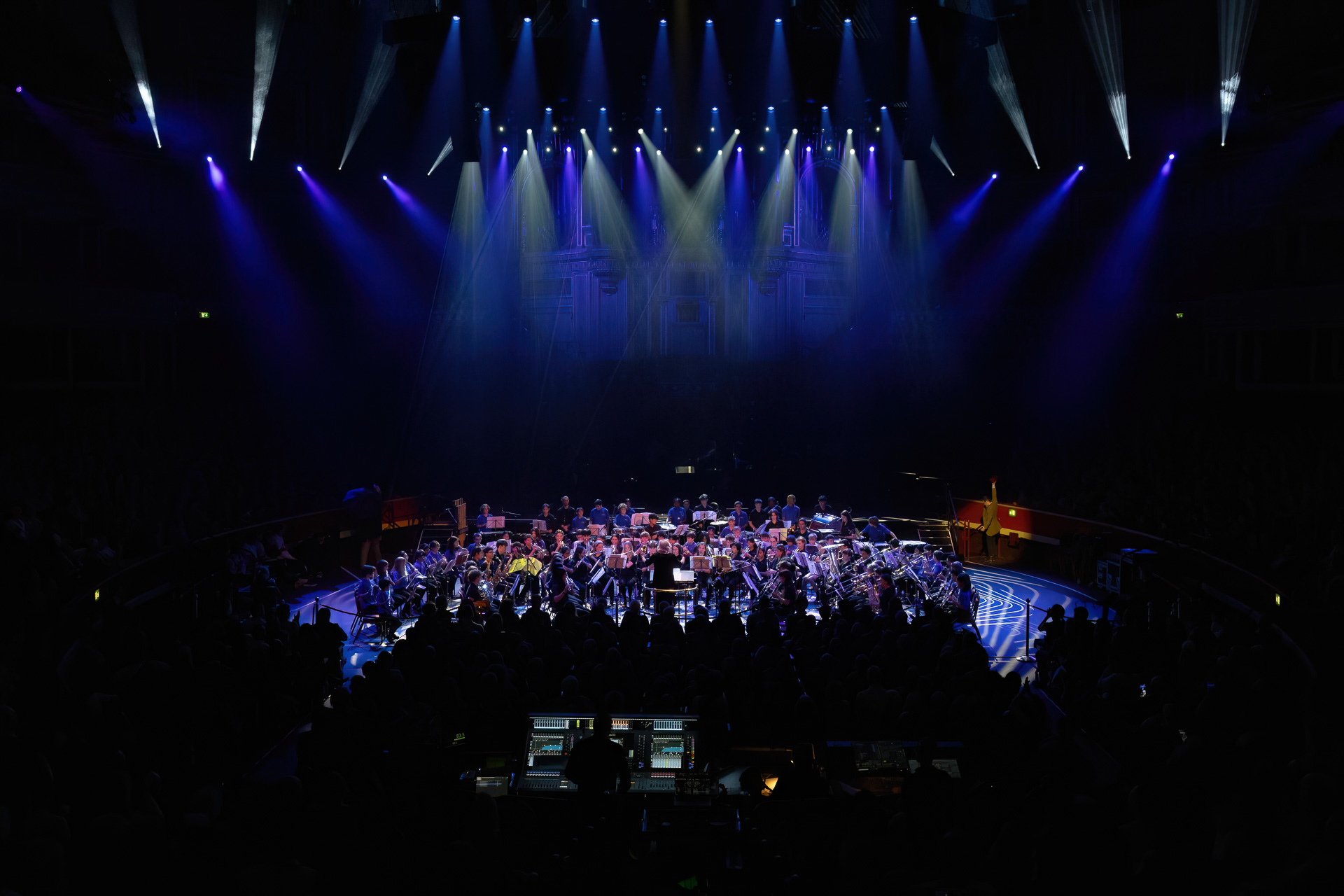 A symphony orchestra performs on a large stage under dramatic blue and purple lighting for Drumathon 2025, with musicians seated in a semi-circle and an audience in the dark foreground.