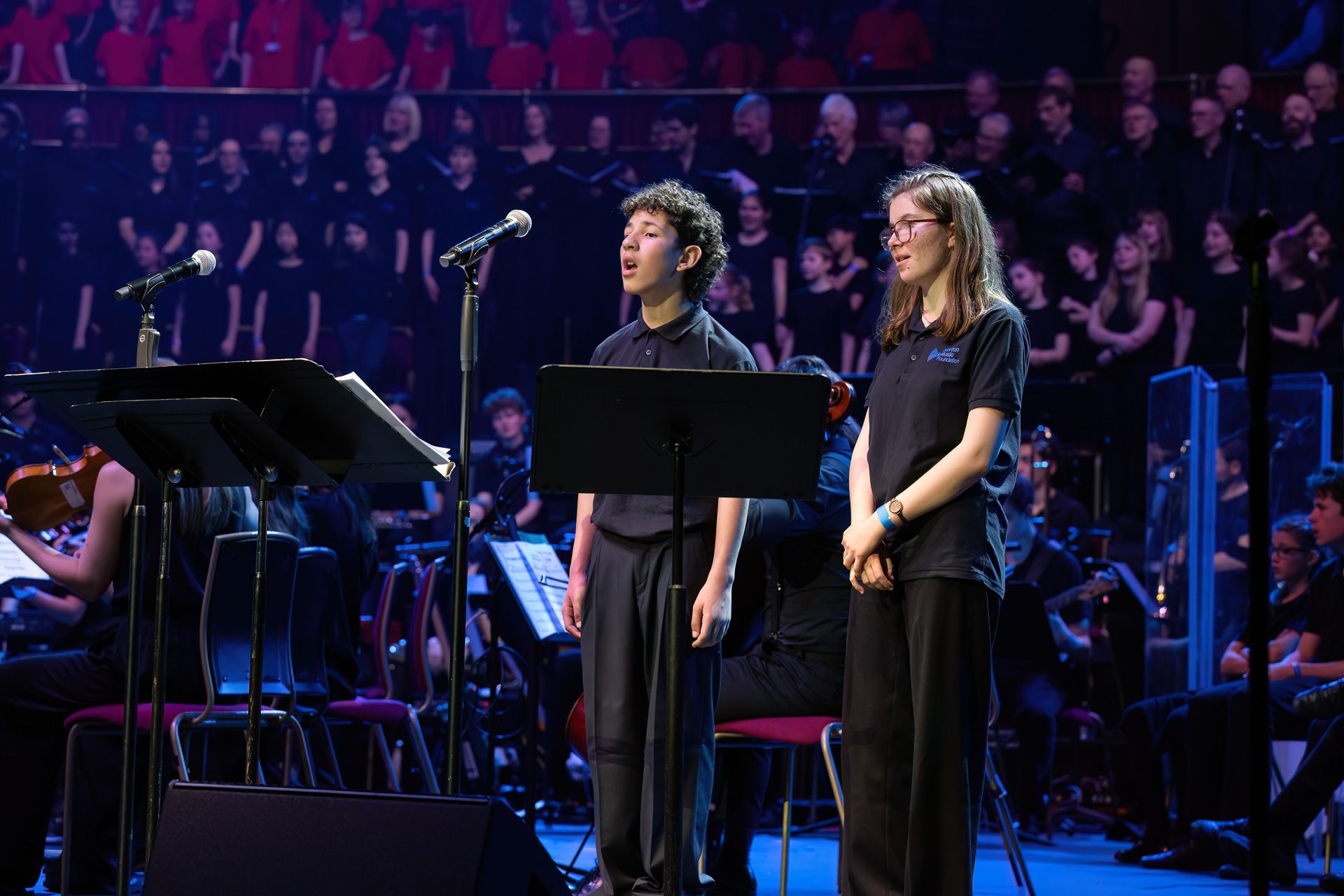 Two young performers stand on stage in front of microphones and music stands, singing with an orchestra and large choir under blue stage lighting at Drumathon 2025.