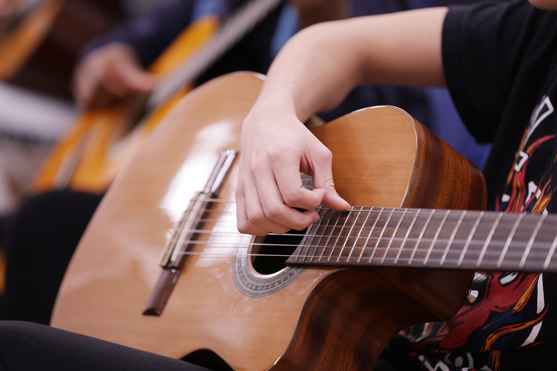 A person wearing a black shirt strums the strings of a classical acoustic guitar, with another person playing a guitar in the blurred background.