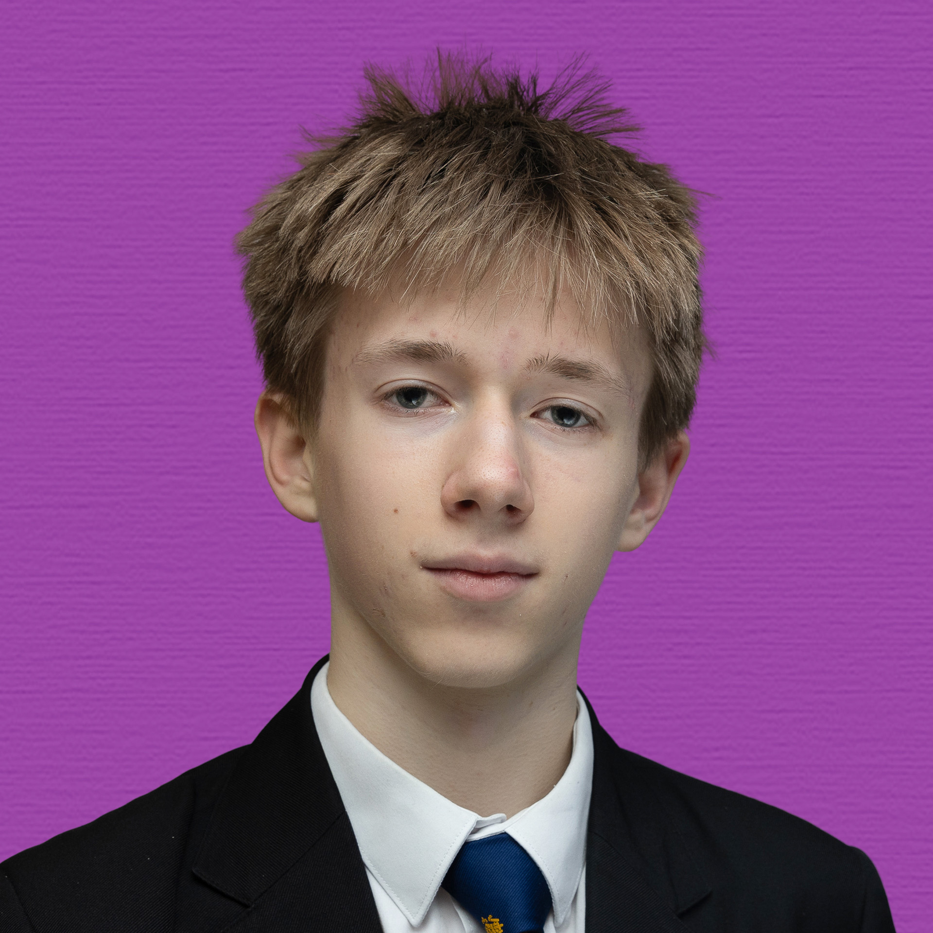 A young person with short, tousled light brown hair wearing a suit, white shirt, and blue tie stands in front of a textured purple background, representing the Youth Council with confidence.