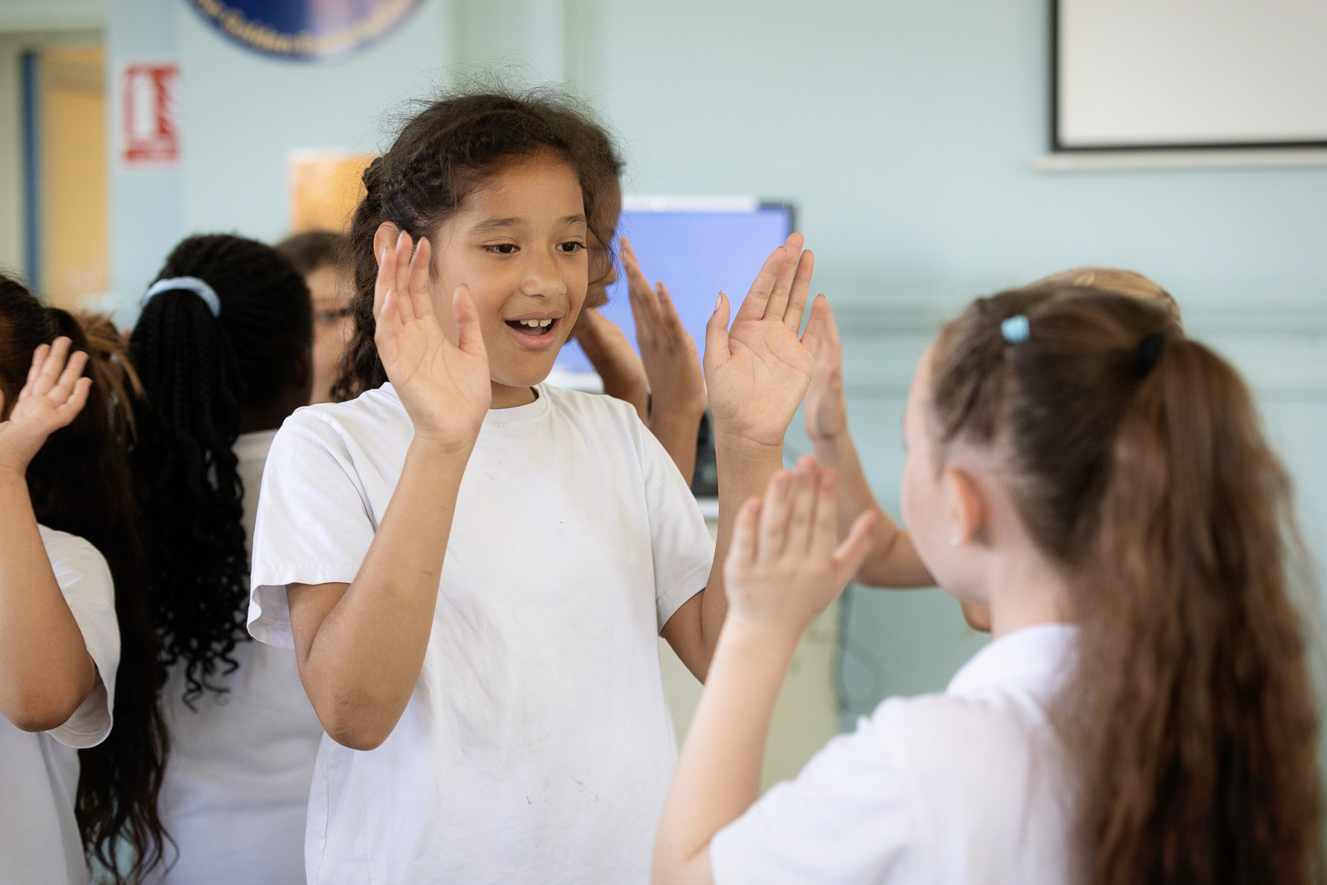 Two young girls in white shirts stand facing each other, smiling and playing a clapping game in a classroom with other children in the background.