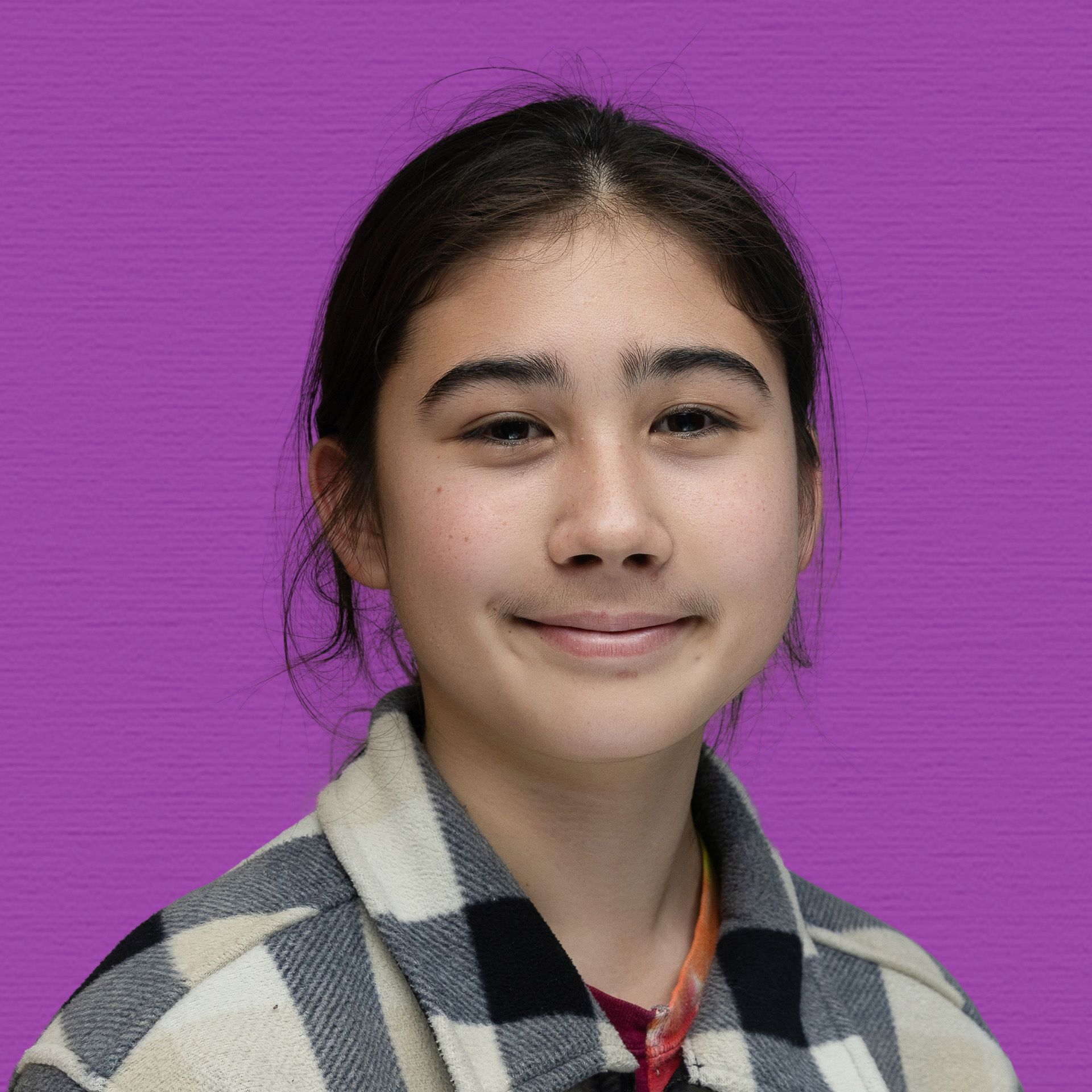 A young person with long dark hair tied back, wearing a black-and-white checked jacket, stands in front of a purple background, smiling gently at the camera—proudly representing the Youth Council.