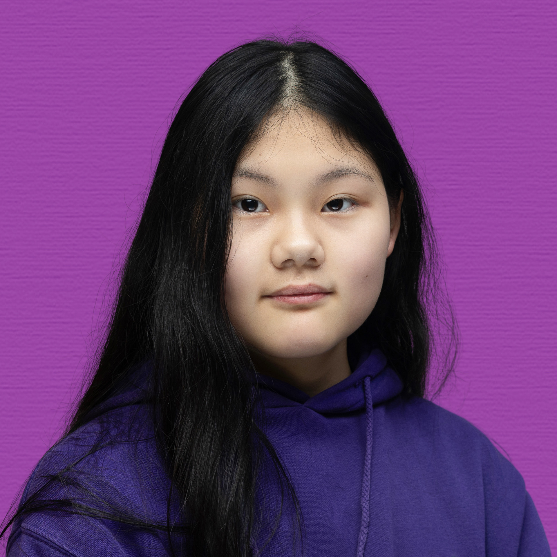 A young girl with long black hair wearing a purple hoodie poses in front of a solid purple background, calmly looking at the camera, representing the spirit of the Youth Council.
